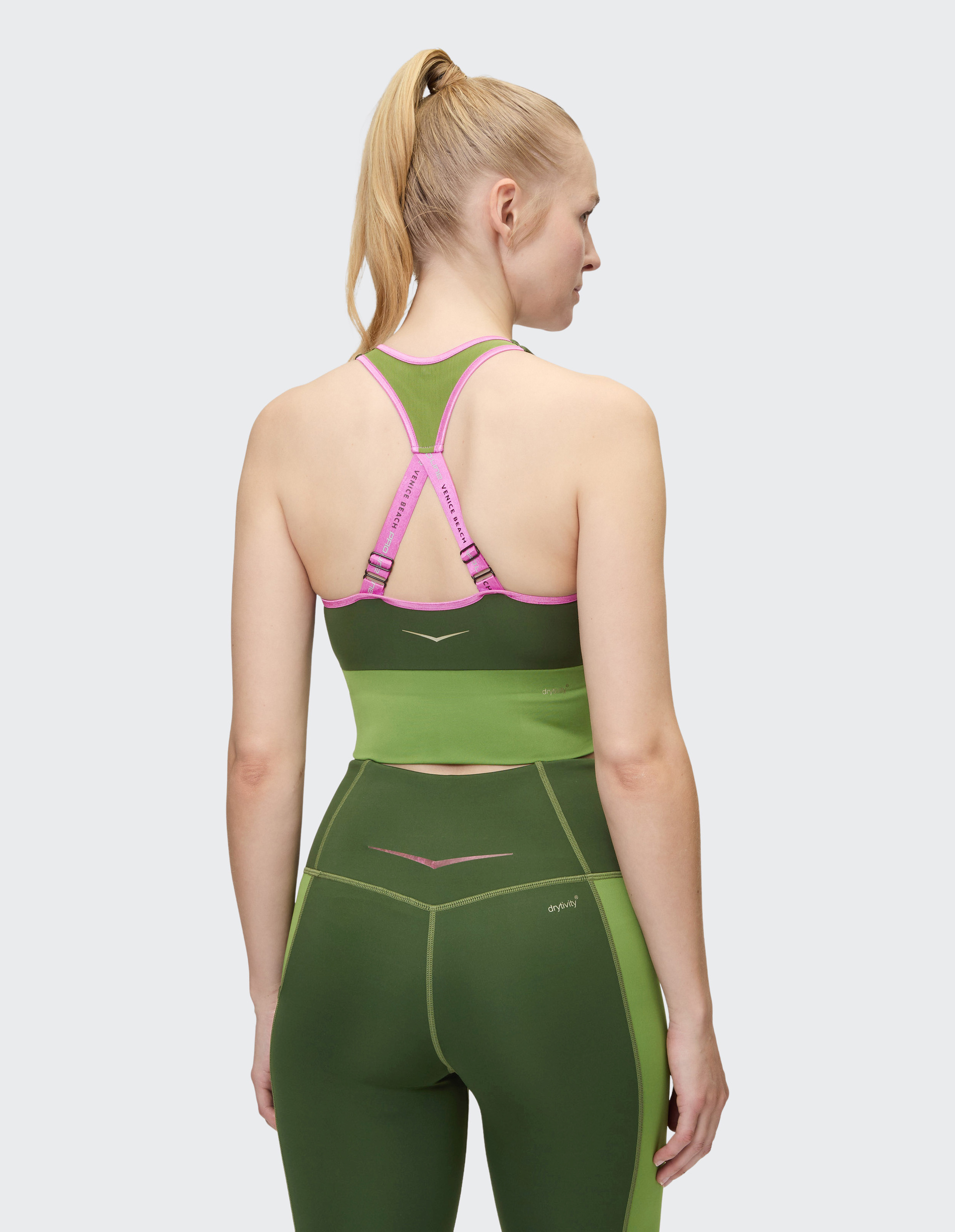 Frau mit blondem Pferdeschwanz trägt grüne Sportbekleidung mit einem rosa, detailreichem Rückenträger auf grauem Hintergrund.
