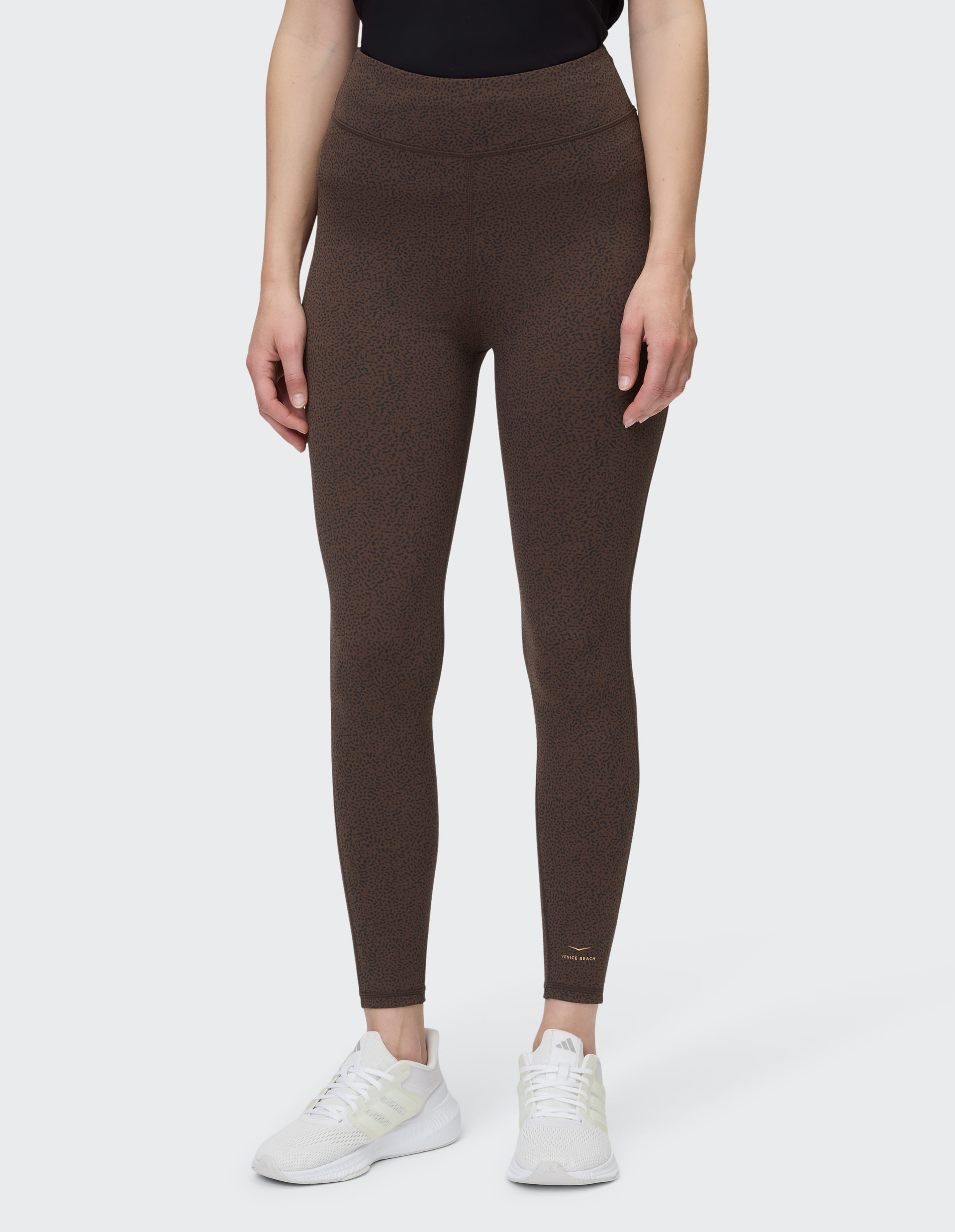 Person trägt eine braune, gemusterte Leggings mit hohem Bund und weiße Sportschuhe, Oberkörper ist teilweise sichtbar.