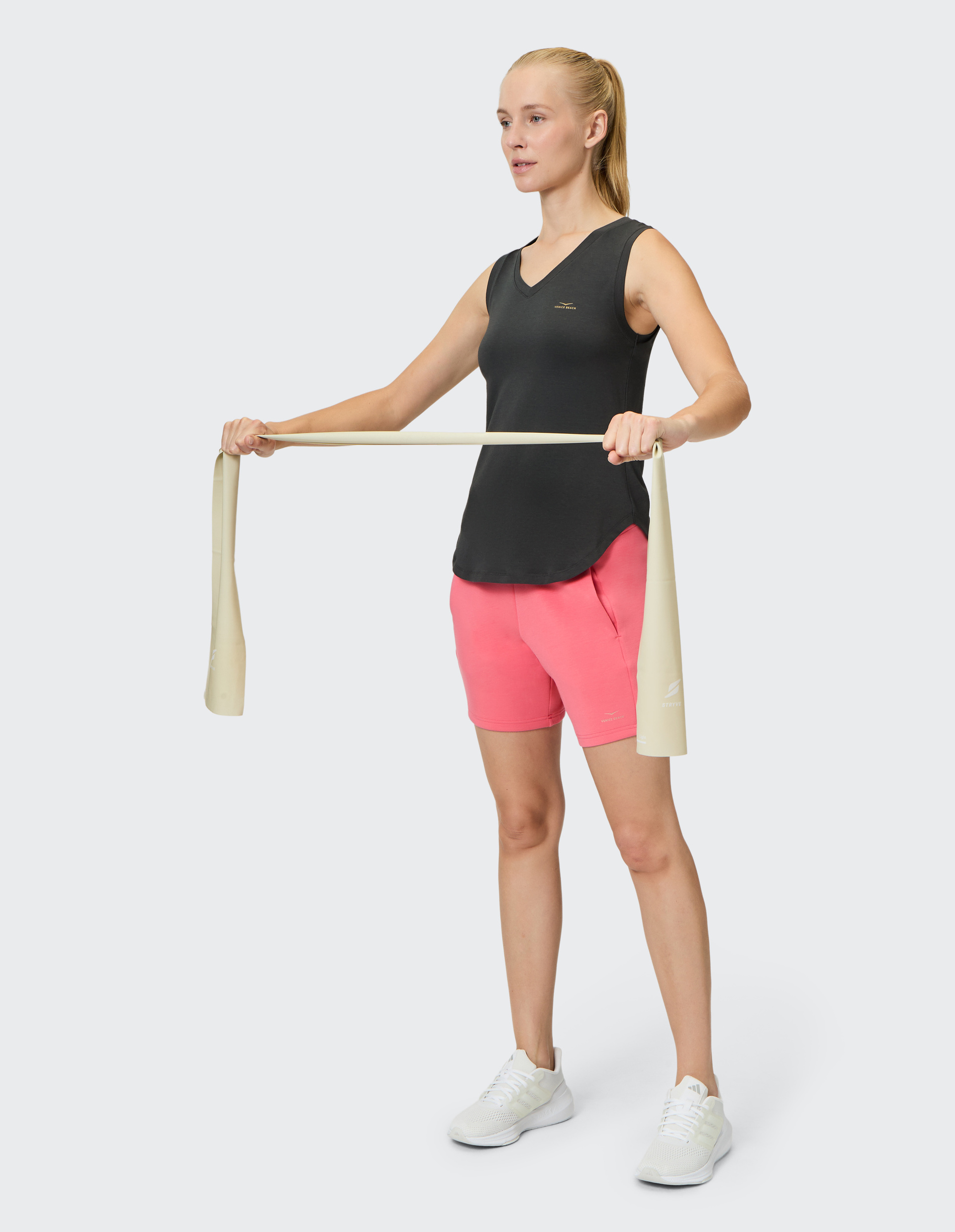 Frau in schwarzem Tanktop und pinken Shorts hält ein Fitnessband, steht aufrecht mit auseinandergezogenen Armen, trägt weiße Turnschuhe.