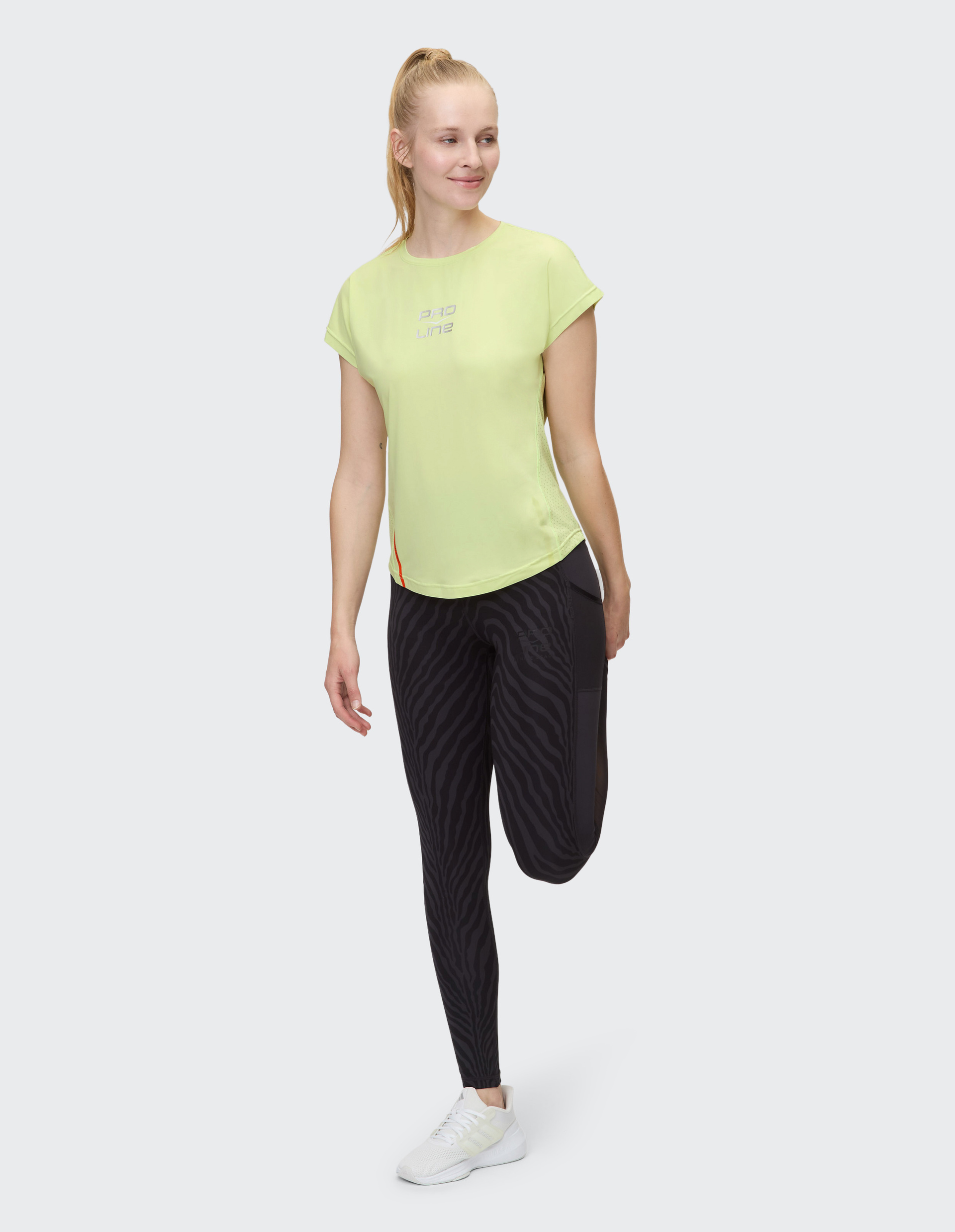Eine Frau in Sportkleidung dehnt ihr Bein, trägt ein gelbes Shirt und schwarze Leggings mit Zebramuster, stehend auf weißem Hintergrund.