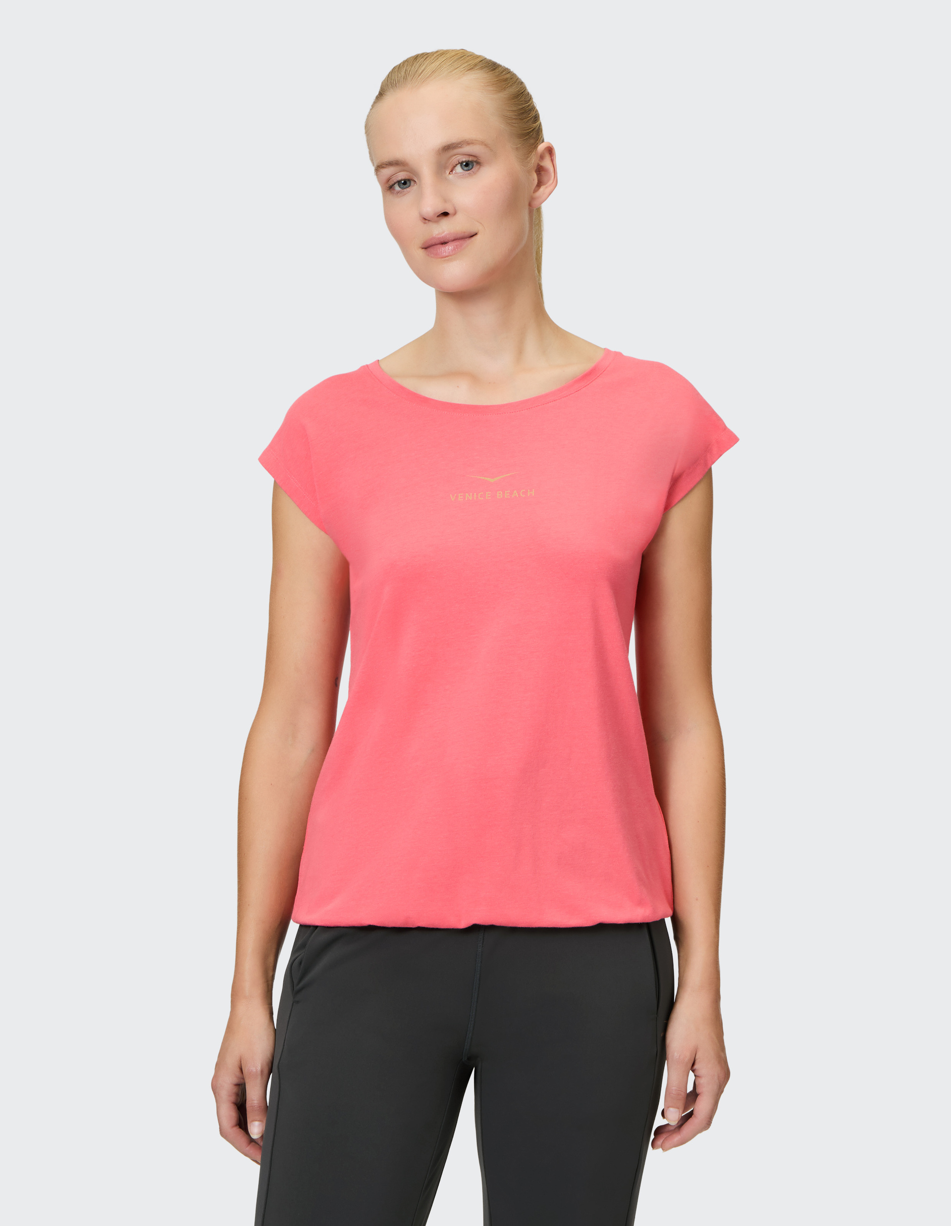 Frau in einem korallenfarbenen T-Shirt mit der Aufschrift Venice Beach und schwarzen Sporthosen vor neutralem Hintergrund.