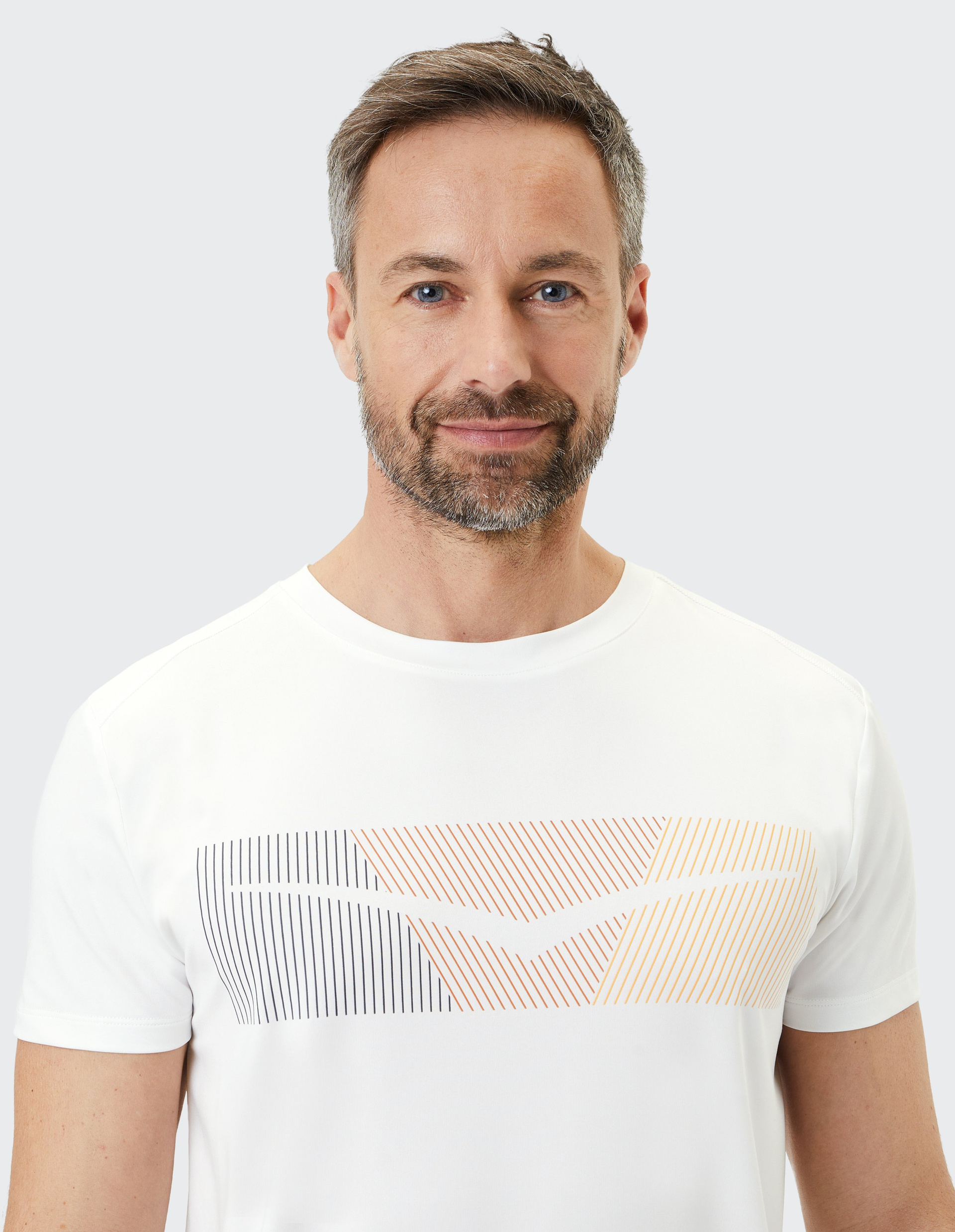 Mann mit kurzem Haar und Bart trägt ein weißes T-Shirt mit abstraktem Linienmuster in Schwarz und Orange, vor grauem Hintergrund.