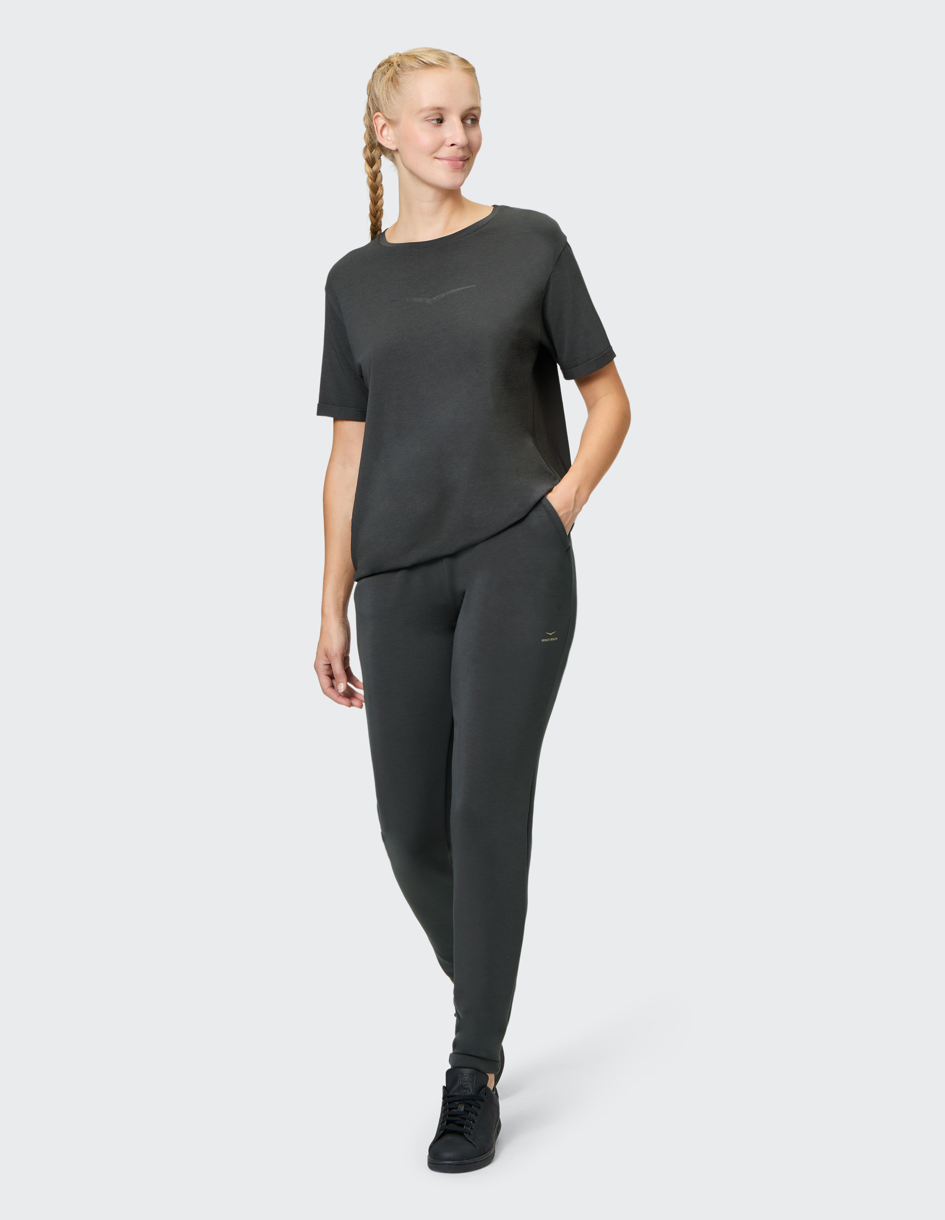 Frau mit seitlich geflochtenem Zopf trägt dunkelgraues T-Shirt und Jogginghose, lächelt und blickt zur Seite; Hände in Hosentaschen.