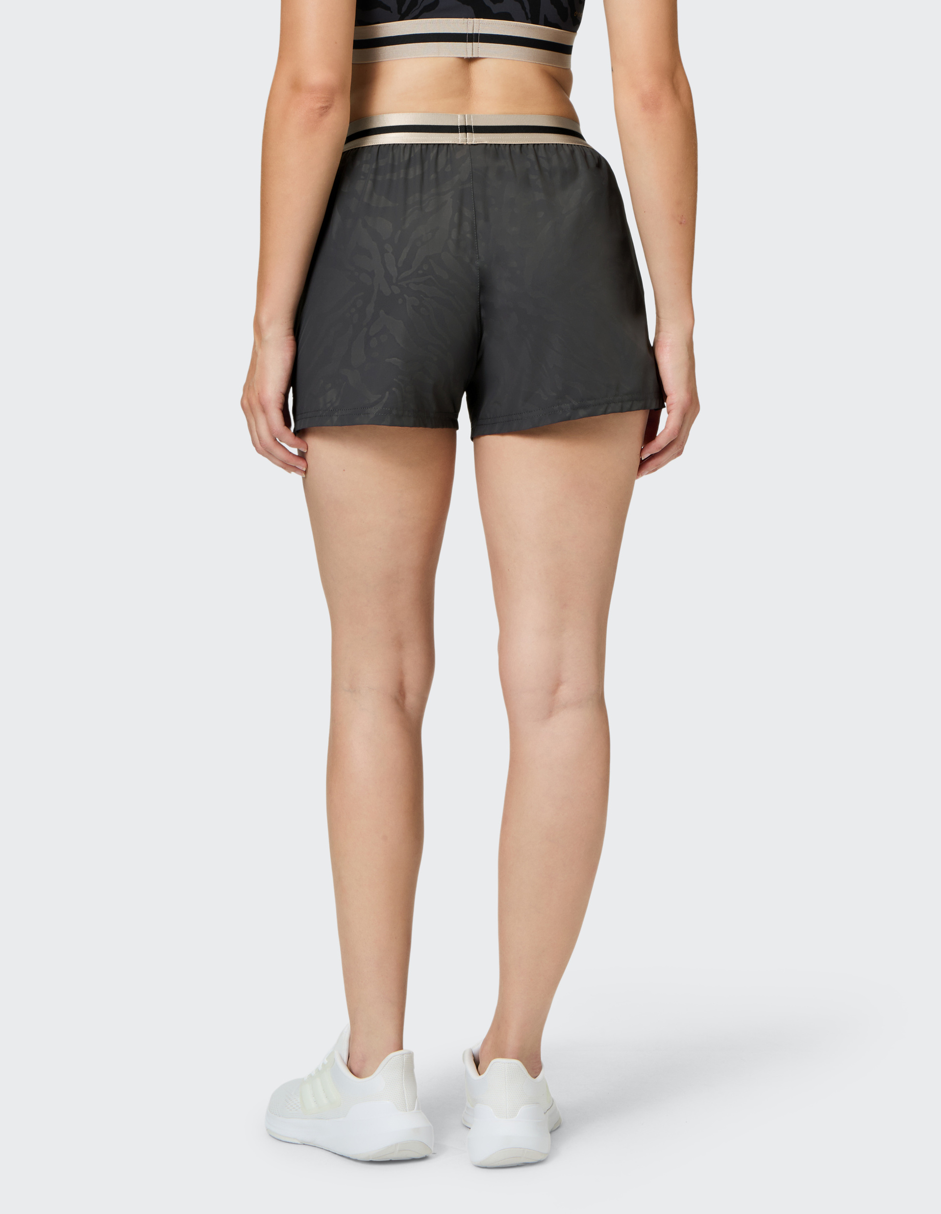 Eine Person steht von hinten, trägt schwarze Shorts, ein passendes Oberteil und weiße Turnschuhe auf grauem Hintergrund.
