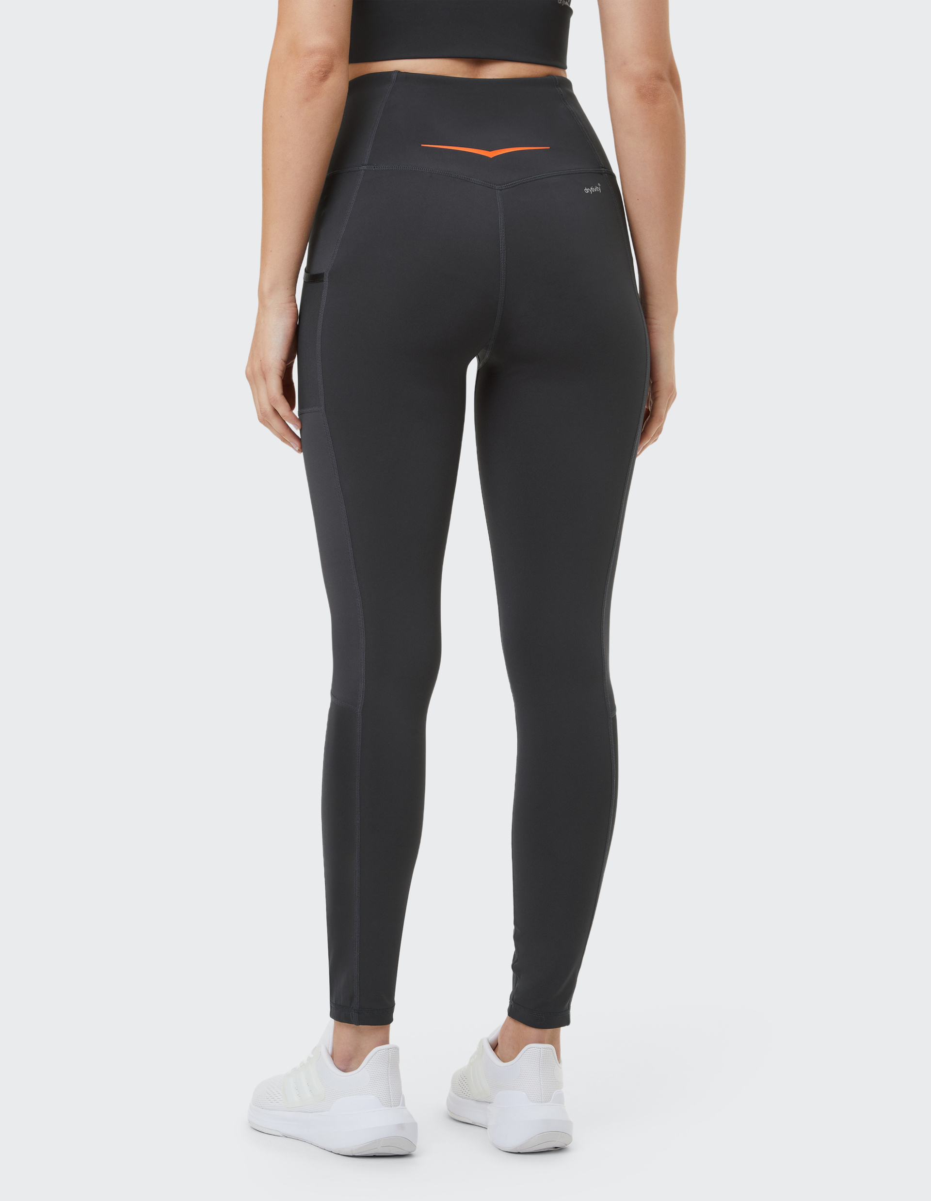 Rückansicht einer Person in dunklen Leggings mit hoher Taille, orange Details, schwarzem Top und weißen Sneakern auf hellem Hintergrund.