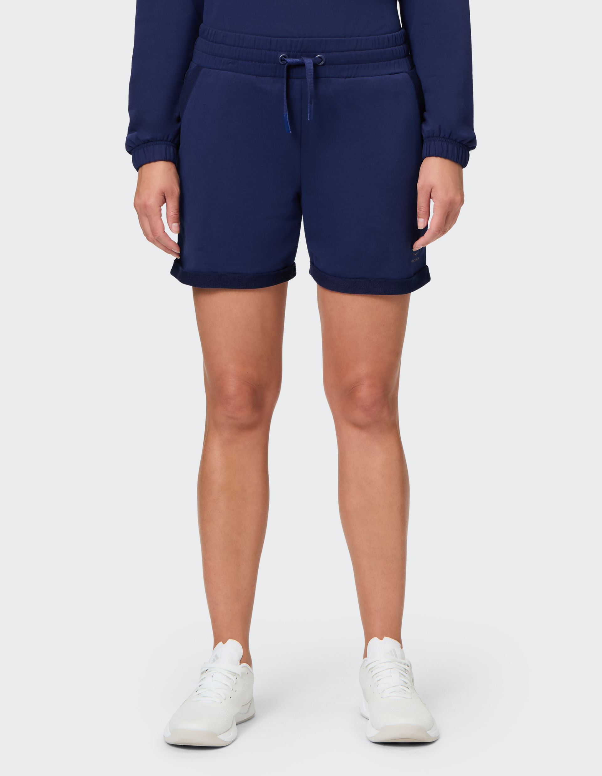 Person trägt dunkelblaue Trainingsshorts mit Tunnelzug und weißen Sneakers, auf hellem Hintergrund, Oberkörper nicht sichtbar.