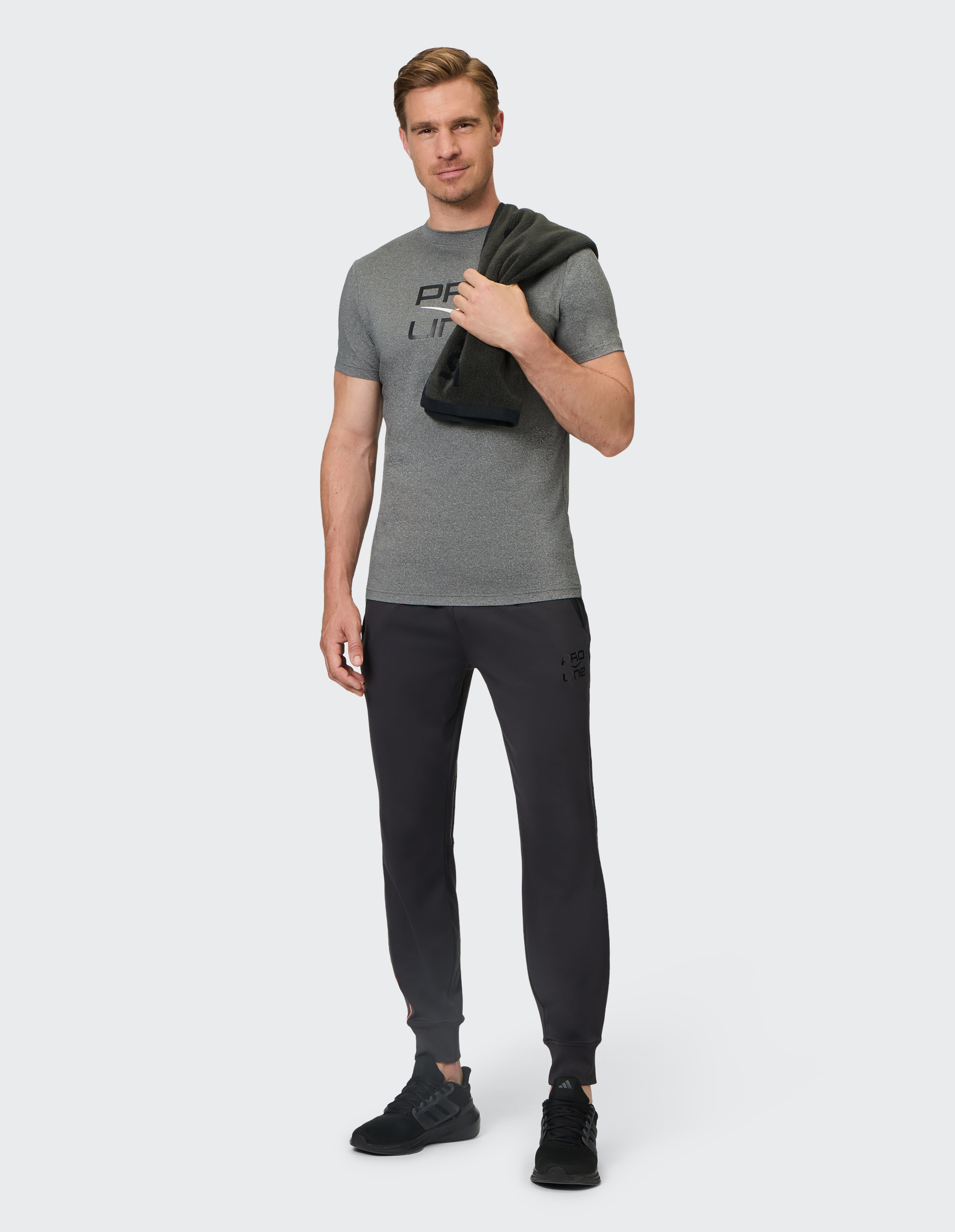 Ein Mann in grauem T-Shirt und dunkler Jogginghose hält ein Handtuch, steht entspannt vor einem neutralen Hintergrund.