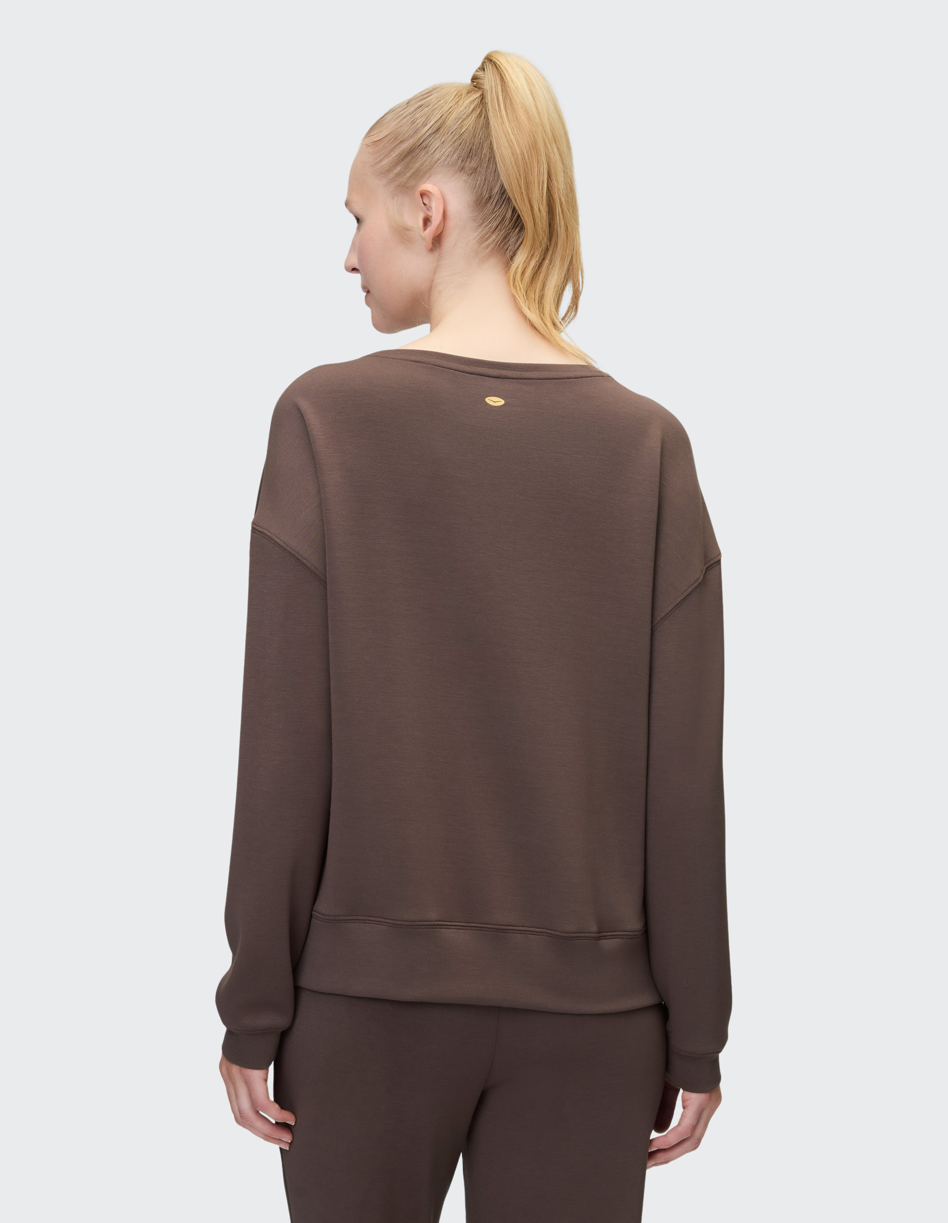 Person mit blondem Pferdeschwanz trägt braunen Pullover und Hose, von hinten fotografiert, auf grauem Hintergrund.