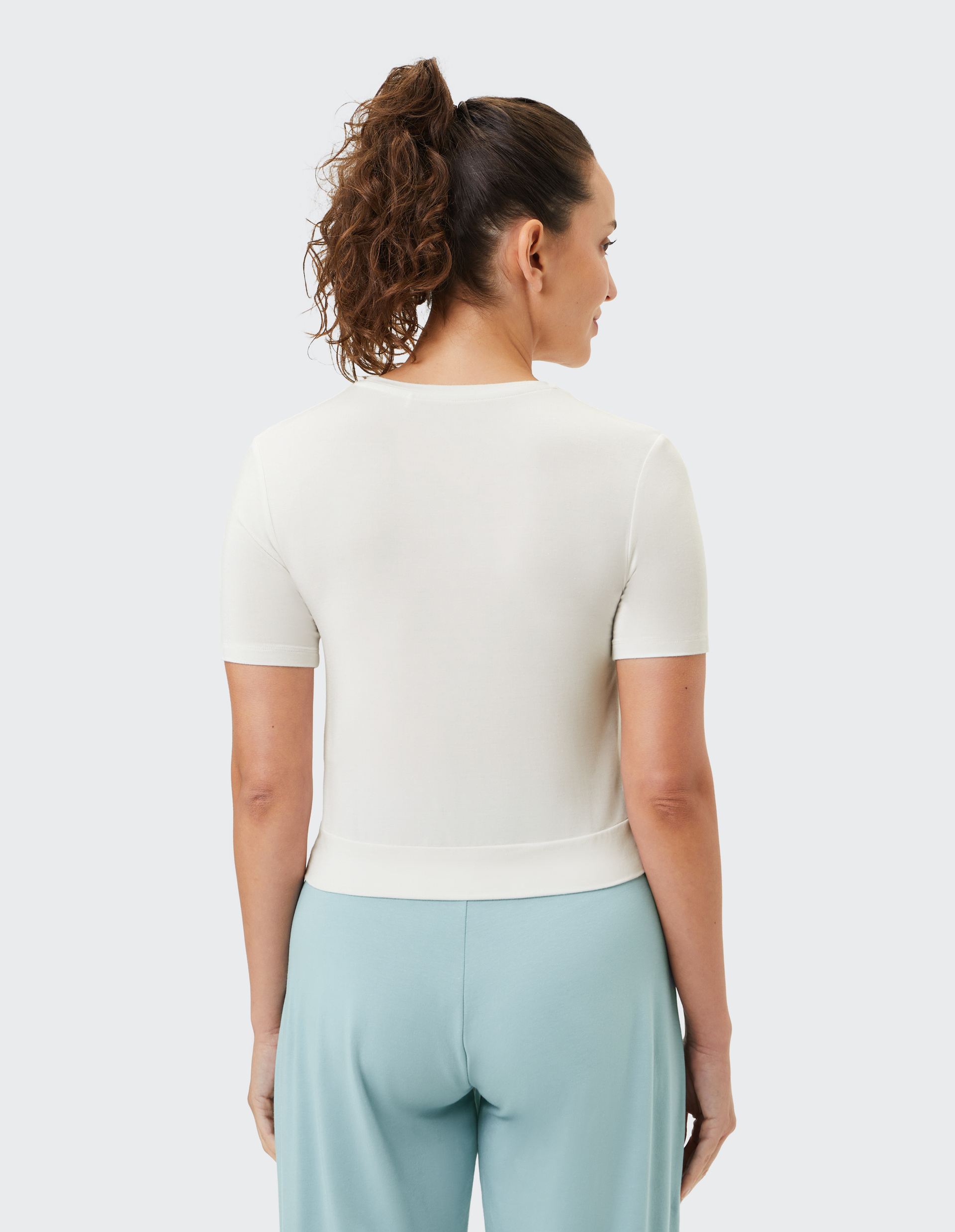 Frau mit lockigem Pferdeschwanz trägt weißes Shirt und hellblaue Hose, von hinten gesehen, vor weißem Hintergrund.