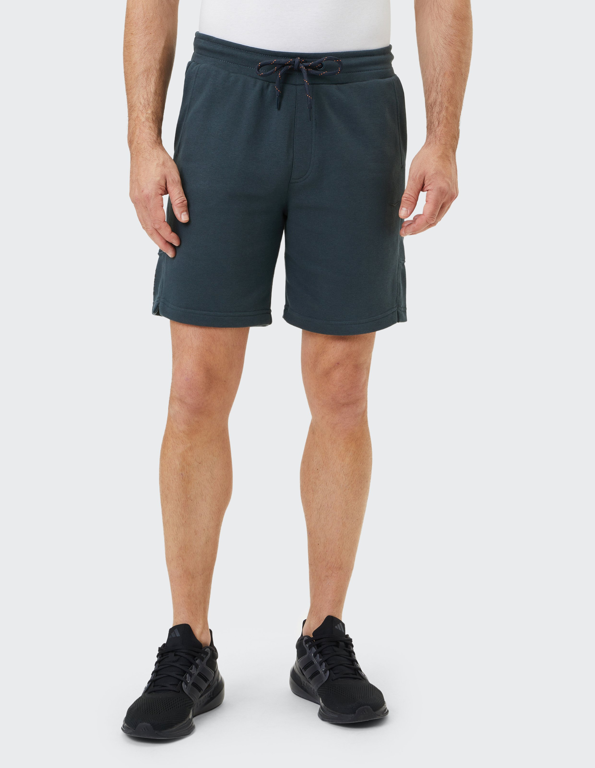 Mann trägt dunkelblaue Shorts und schwarze Sneaker auf hellem Hintergrund, die Arme locker an den Seiten, T-Shirt nicht sichtbar.