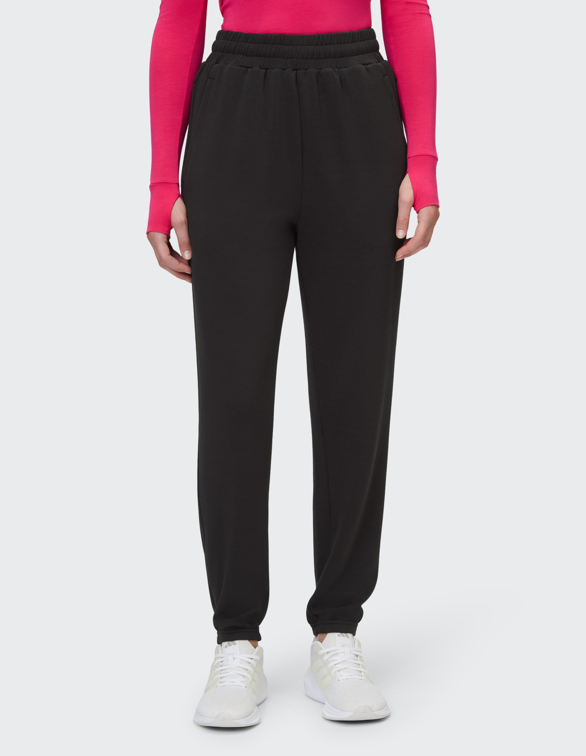 Person in schwarzer Jogginghose und weißem Sneaker, trägt ein langärmliges, pinkes Oberteil, hellgrauer Hintergrund.