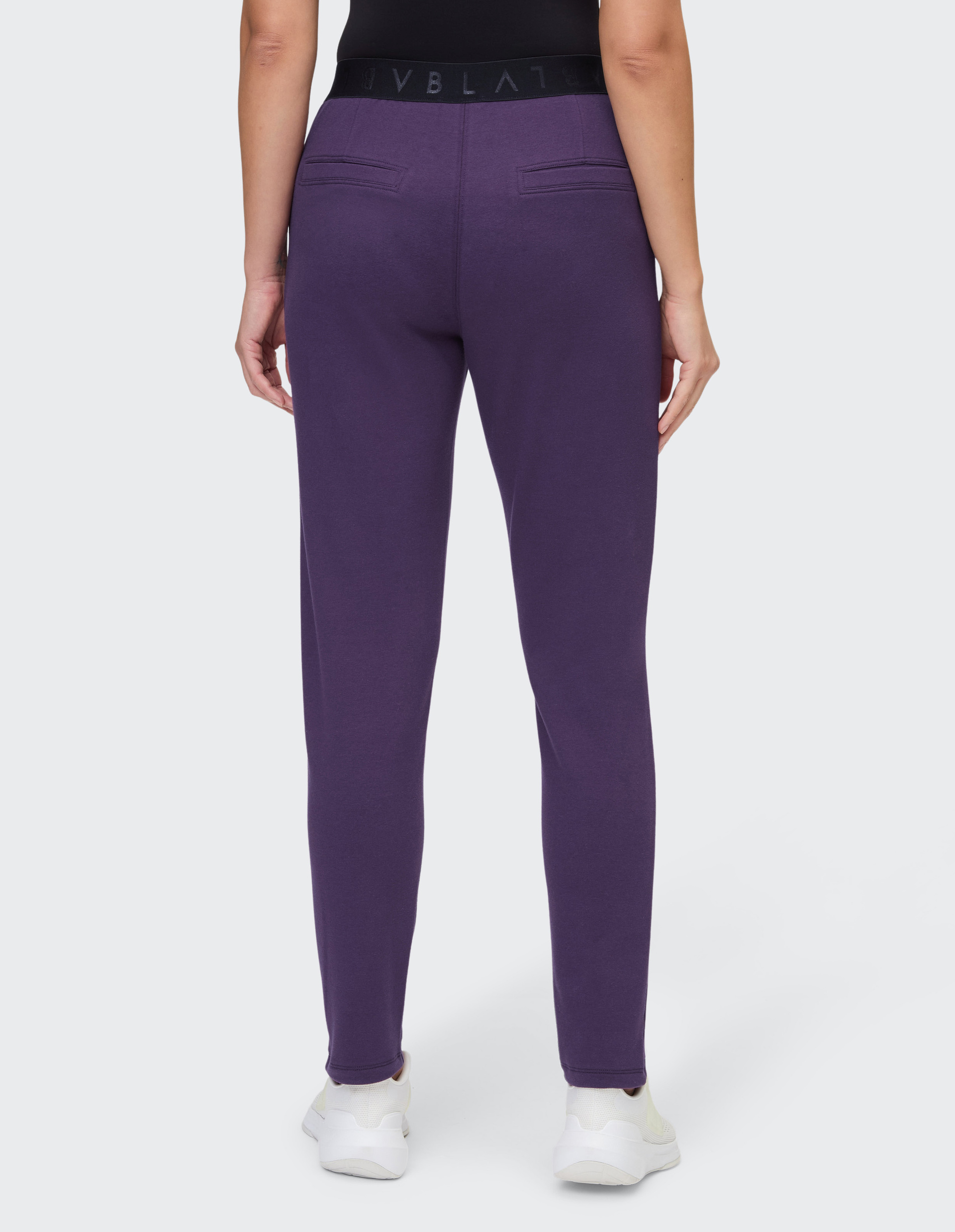 Person trägt lila Jogginghose mit schwarzem Bund und weißen Sneakers, fotografiert von hinten vor weißem Hintergrund.