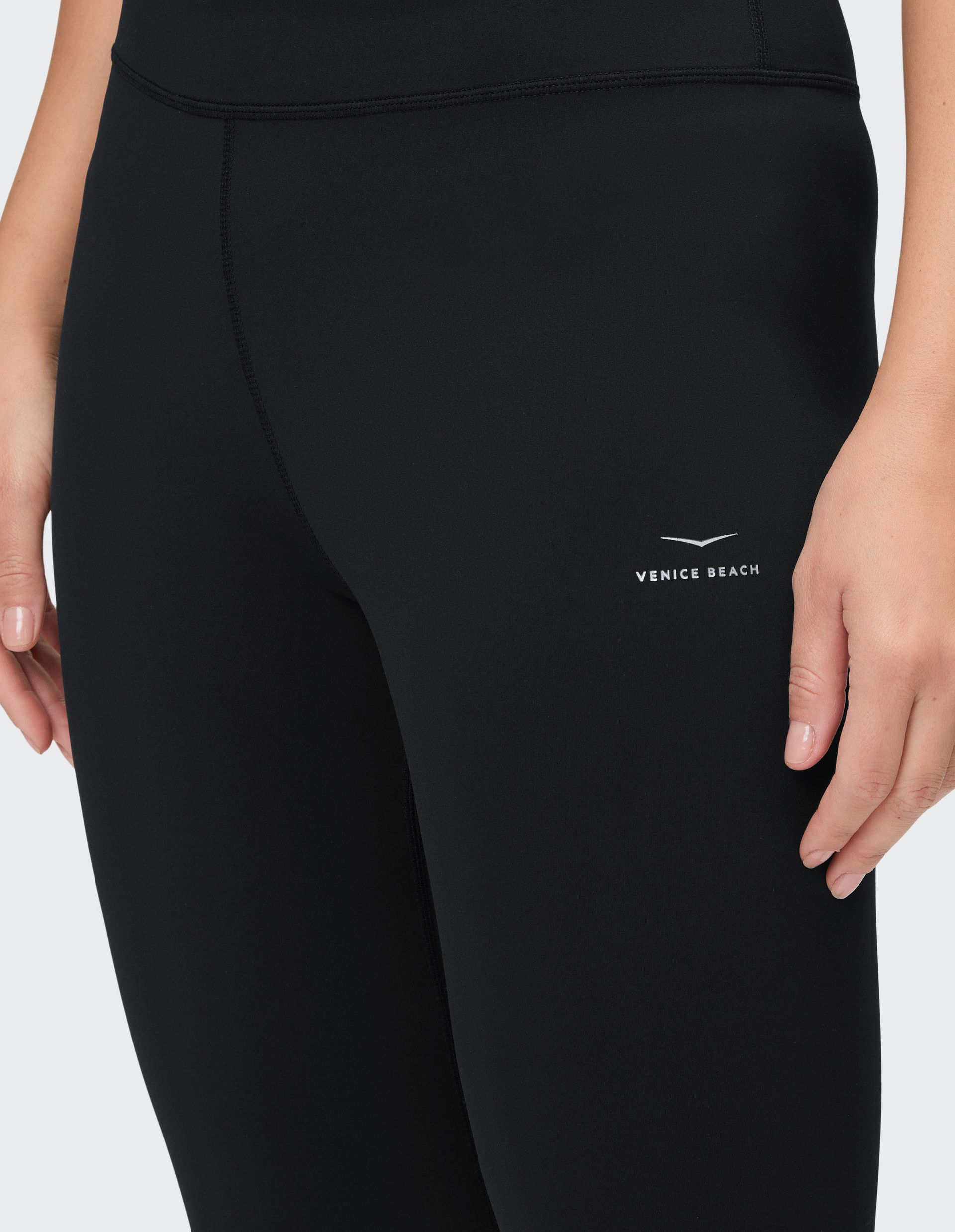 Nahaufnahme einer Person in schwarzer Sportleggings mit Venice Beach-Logo auf dem Oberschenkel und die rechte Hand seitlich sichtbar.