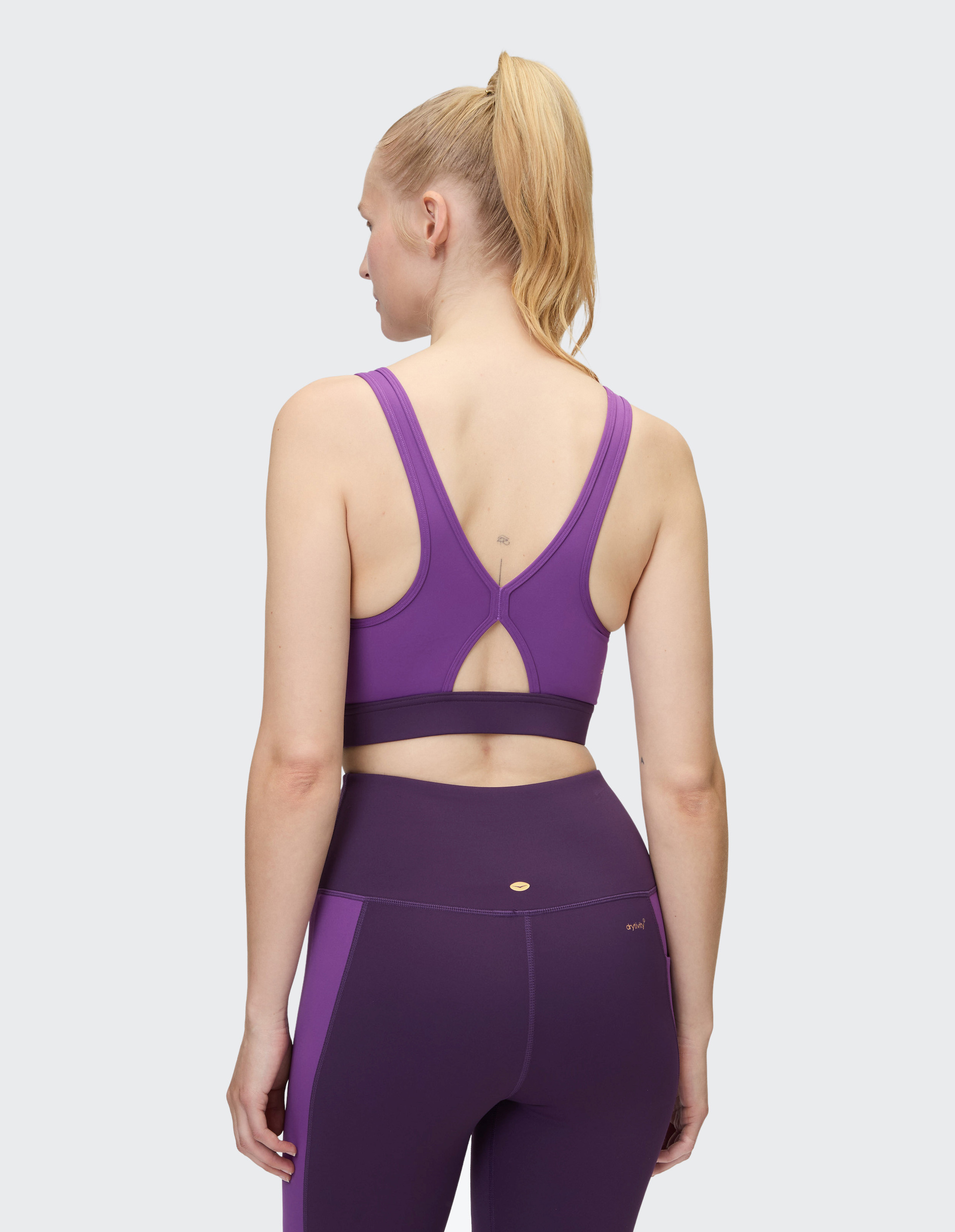 Eine Frau mit blondem Pferdeschwanz trägt ein violettes Sport-BH-Oberteil und passende Leggings, von hinten gesehen.