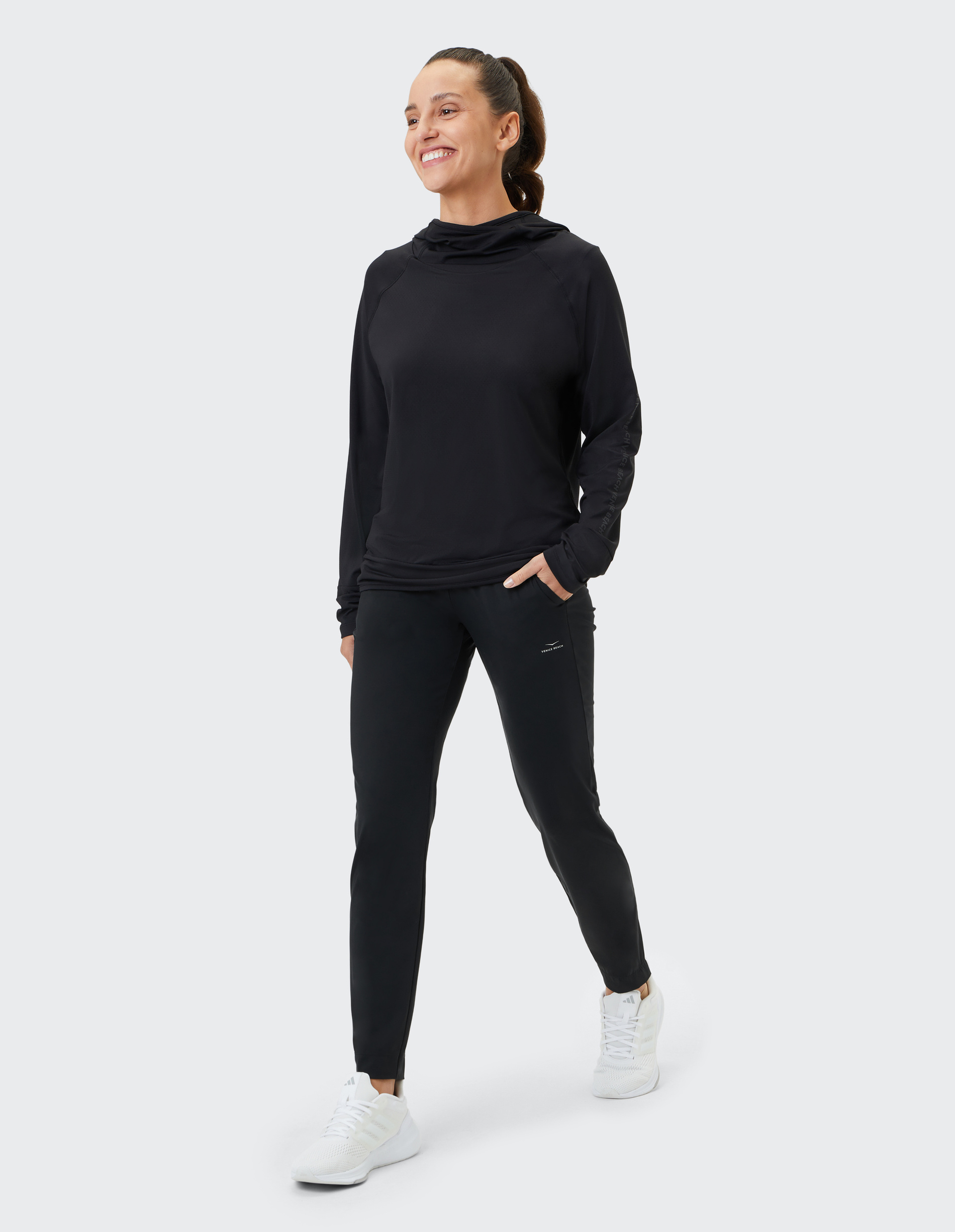 Eine Frau in schwarzem Sportpullover und Jogginghose geht lächelnd, mit einer Hand in der Hosentasche und weißen Sneakers.