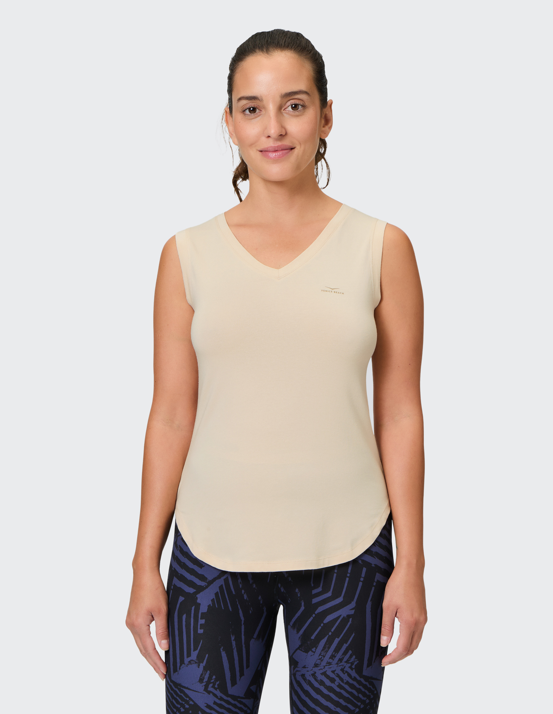 Frau in beige ärmellosem Top und blau-schwarz gemusterten Leggings steht vor neutralem Hintergrund und lächelt.