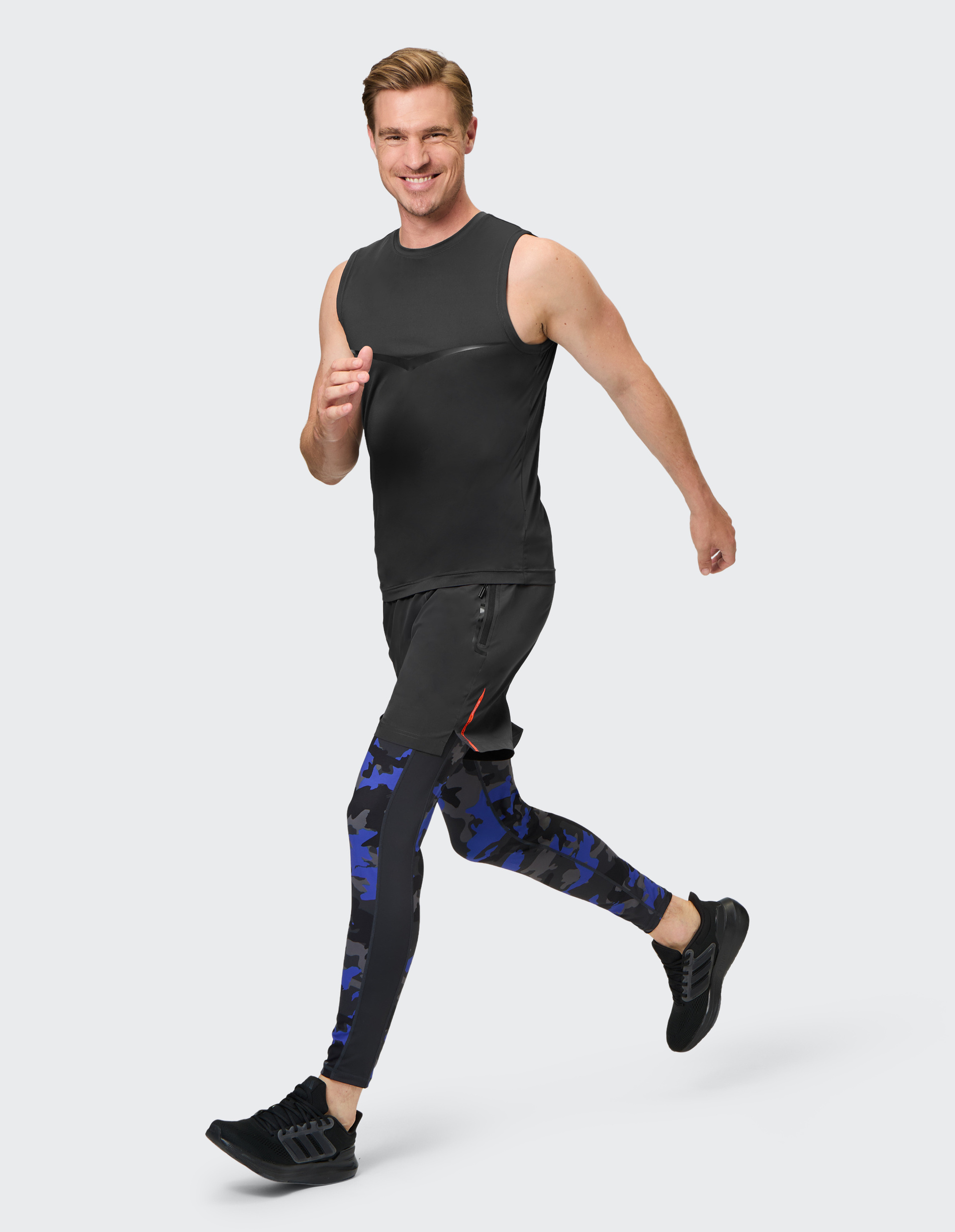 Ein lächelnder Mann joggt in Sportkleidung, bestehend aus schwarzem Tanktop, Shorts und blau-gemusterten Leggings.