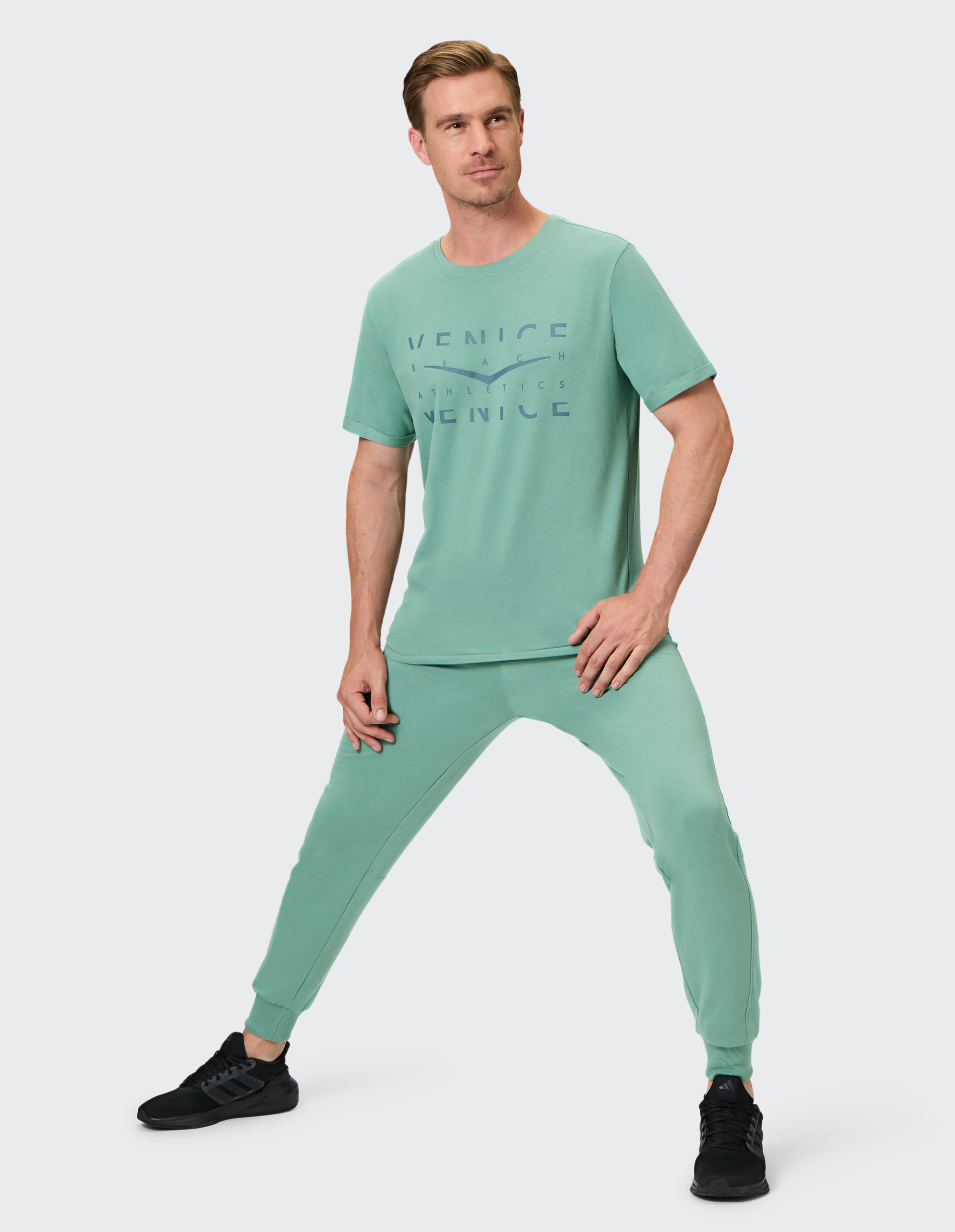 Ein Mann trägt ein mintgrünes Shirt mit "Venice Beach" und passende Jogginghose, posiert leicht nach links gewandt, auf hellem Hintergrund.