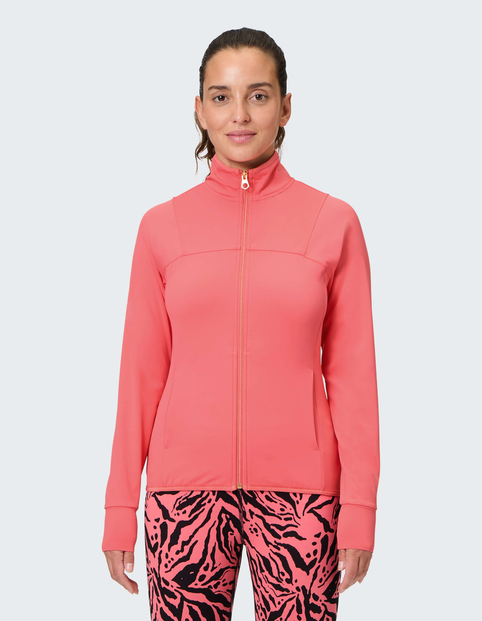 Frau in korallfarbener Jacke mit Reißverschluss und gemusterten, pink-schwarz karierten Leggings vor neutralem Hintergrund.