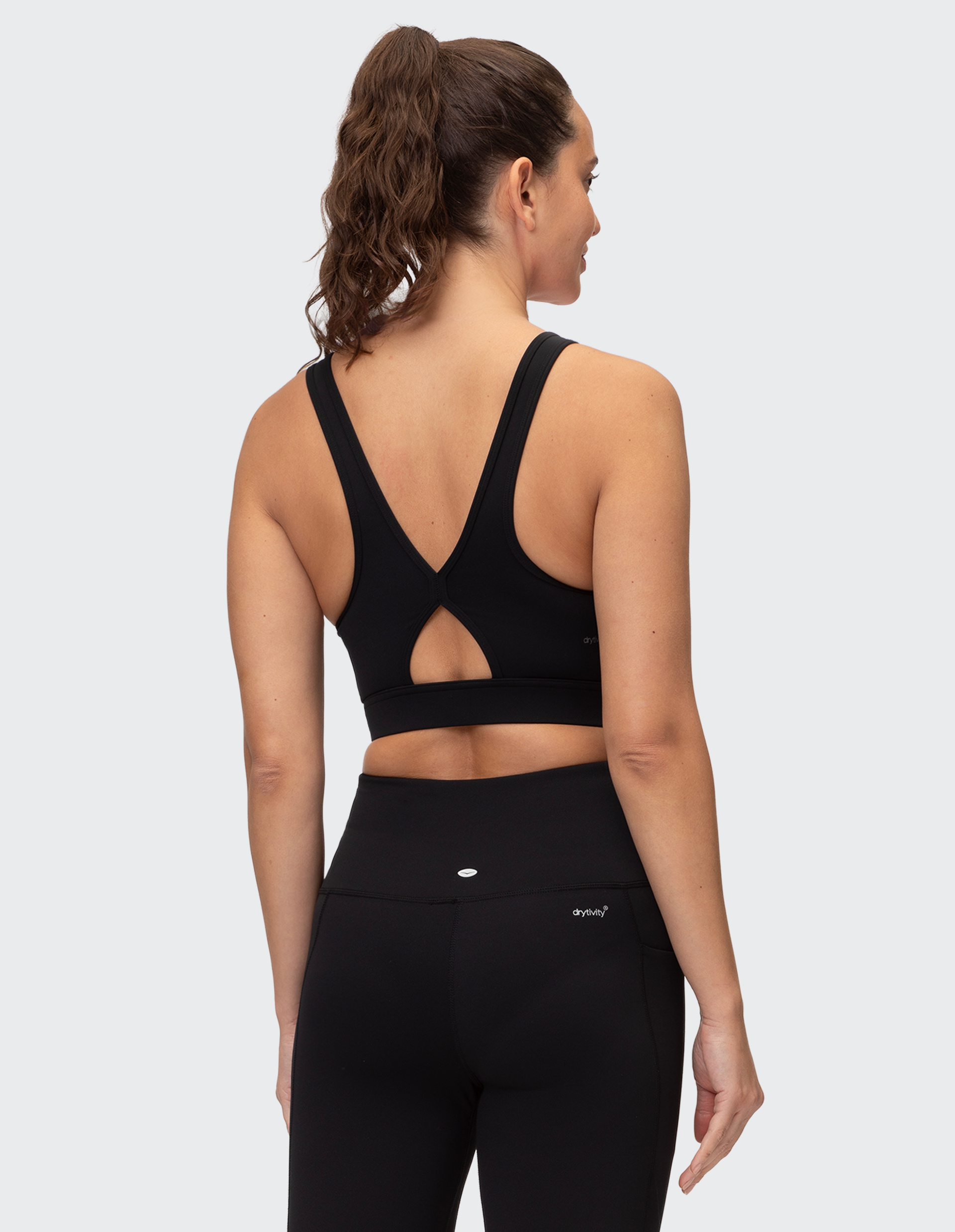 Frau mit dunklem Pferdeschwanz von hinten, trägt ein schwarzes Sporttop mit Rückenausschnitt und passende Leggings vor grauem Hintergrund.