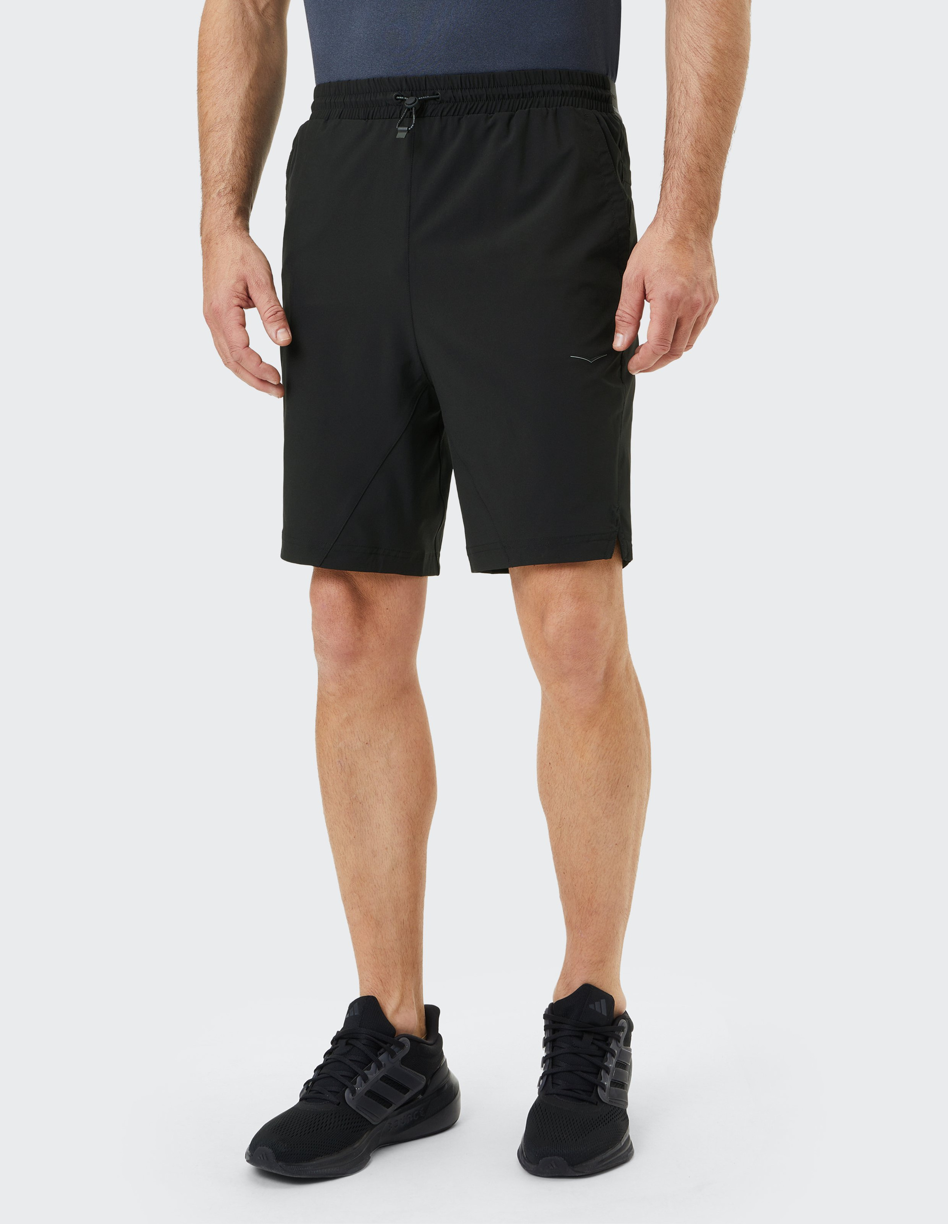 Mann in schwarzem Sportshirt und Shorts, trägt schwarze Turnschuhe; steht aufrecht vor weißem Hintergrund, Hände an den Seiten.