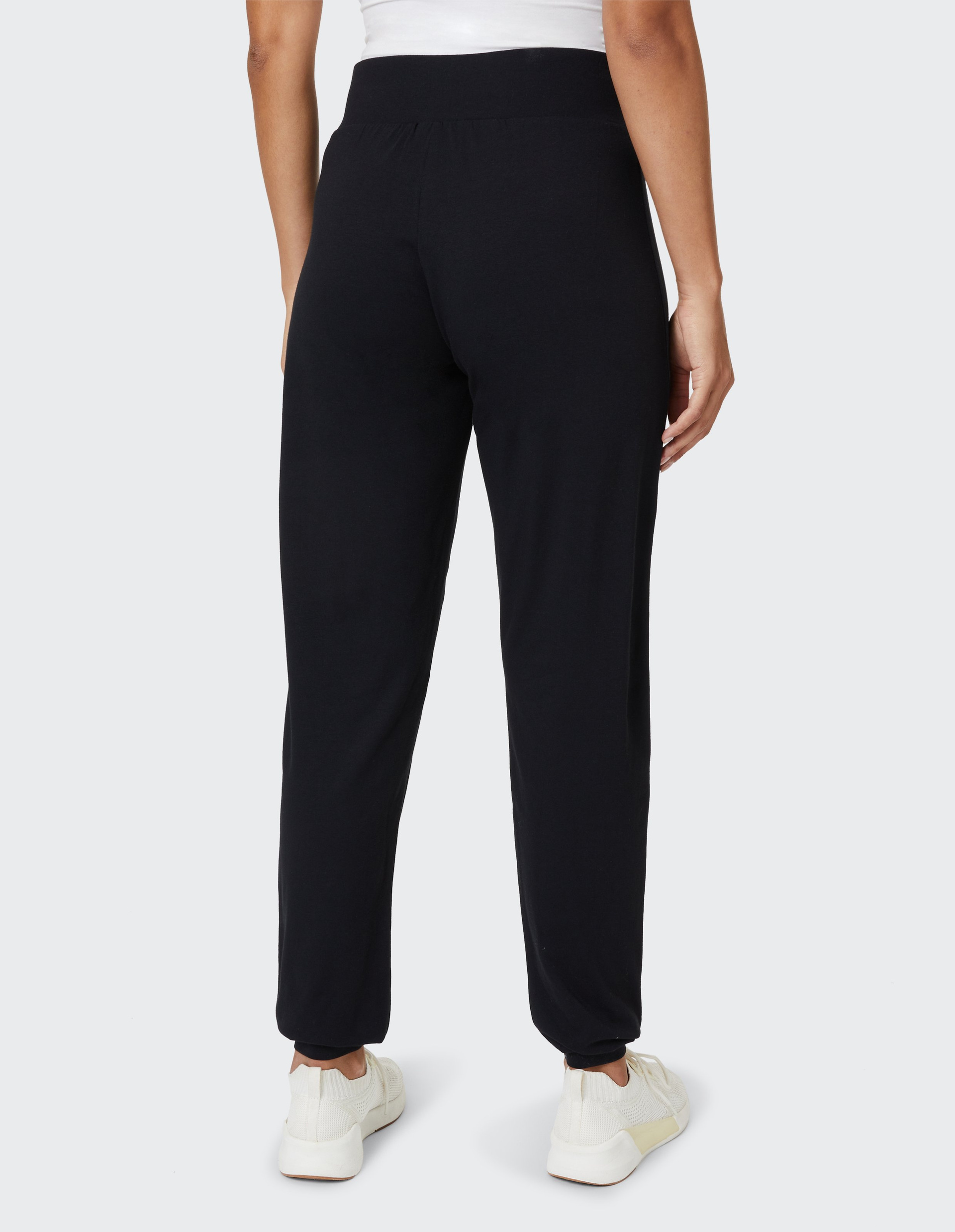 Person von hinten in schwarzer Hose und weißen Turnschuhen, vor grauem Hintergrund, trägt ein weißes Oberteil.