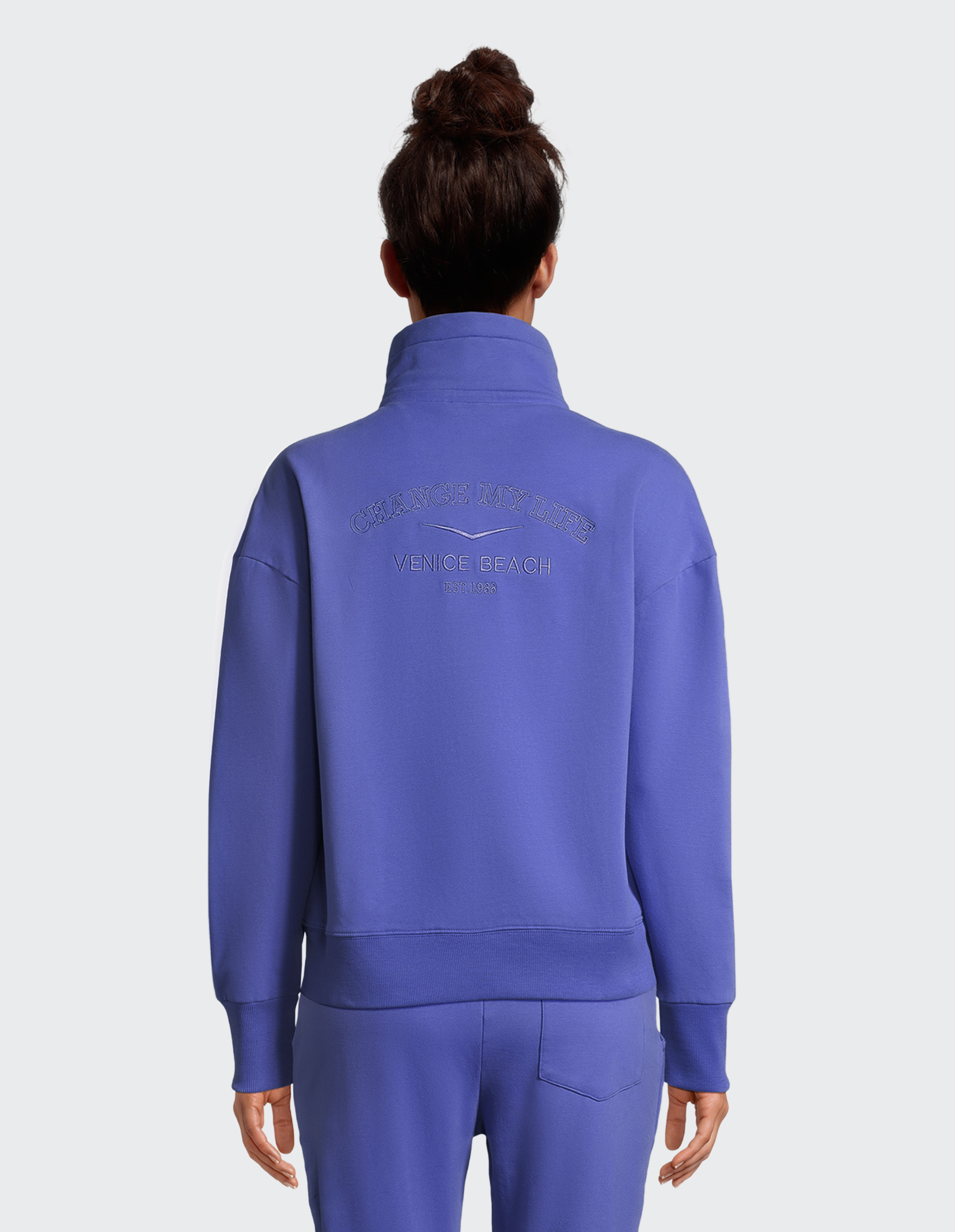 Person in blauem Jogginganzug von hinten, mit hochgestecktem Haar; Rückenaufschrift: Change My Life, Venice Beach 1951-1963.