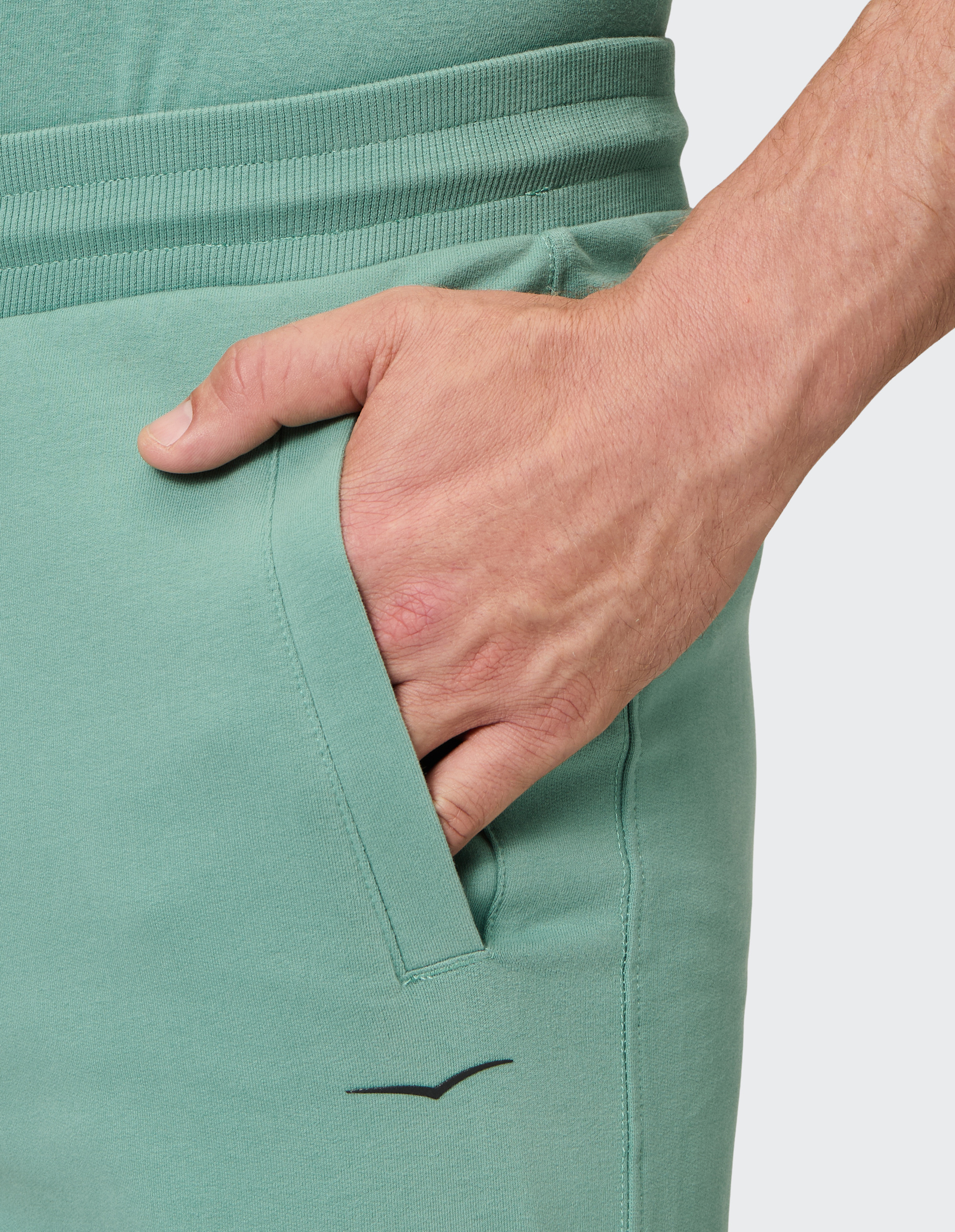 Nahaufnahme einer Hand, die in der Tasche einer mintgrünen Jogginghose steckt, mit geripptem Bund und schwarzem Logo.