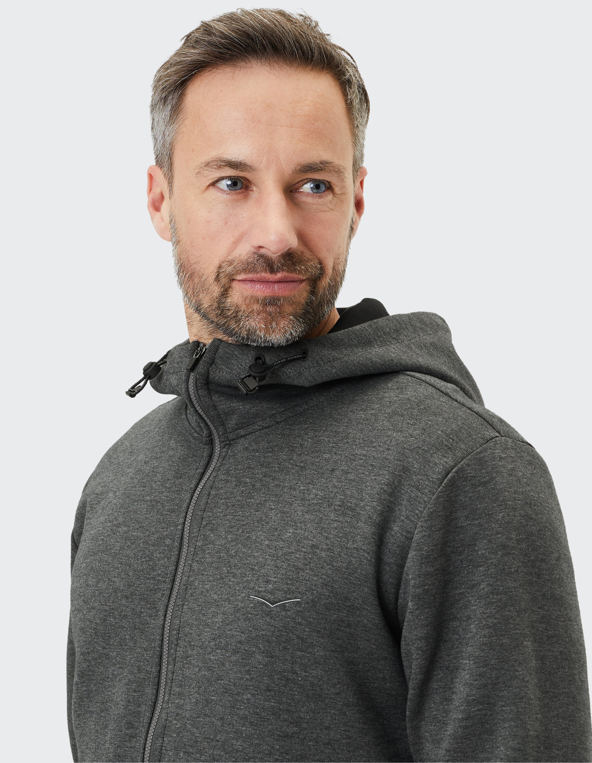 Mann mit kurzem Haar und Bart trägt dunkelgrauen Kapuzenpullover mit Reißverschluss und Logostickerei, blickt nach links.