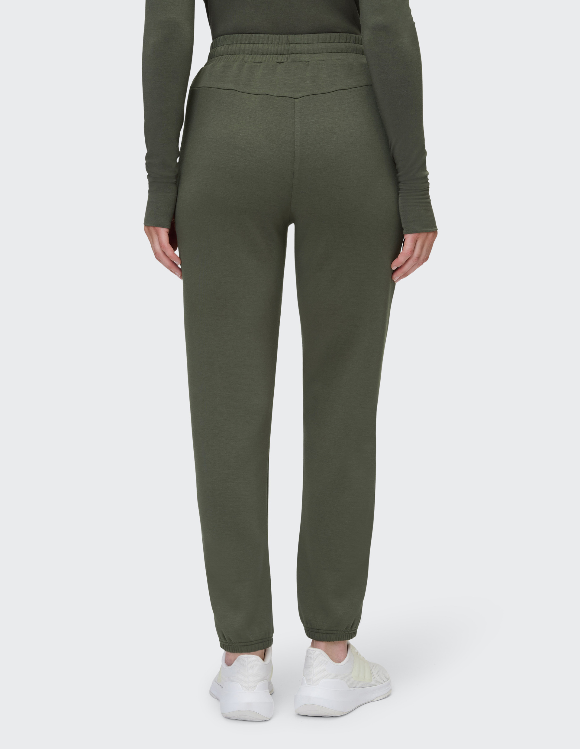 Rückansicht einer Person in einer olivgrünen Jogginghose und weißen Sneakern, die auf hellem Hintergrund steht.