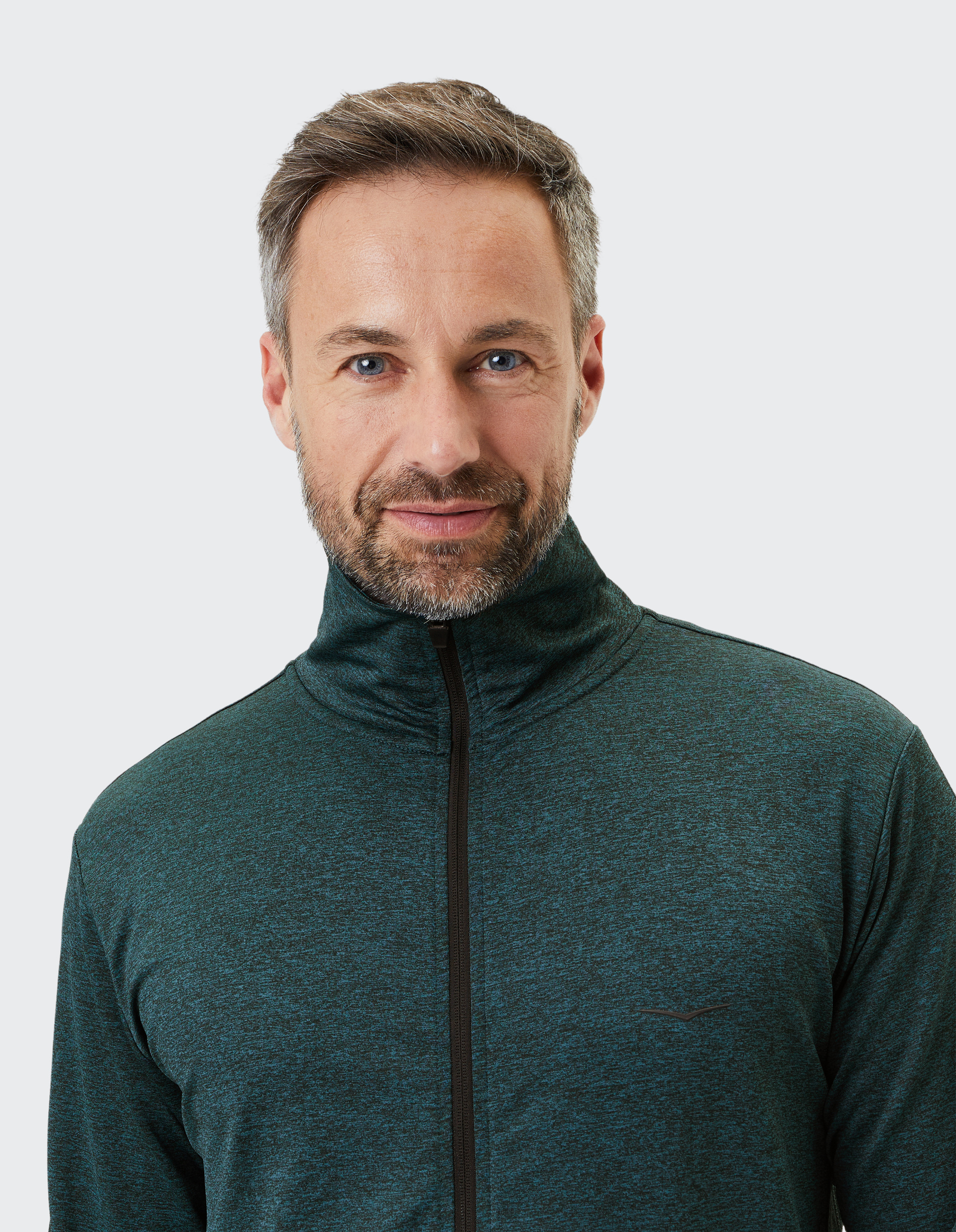 Ein Mann mit braunen Haaren und Bart trägt einen dunkelgrünen Reißverschluss-Pullover vor hellem Hintergrund.