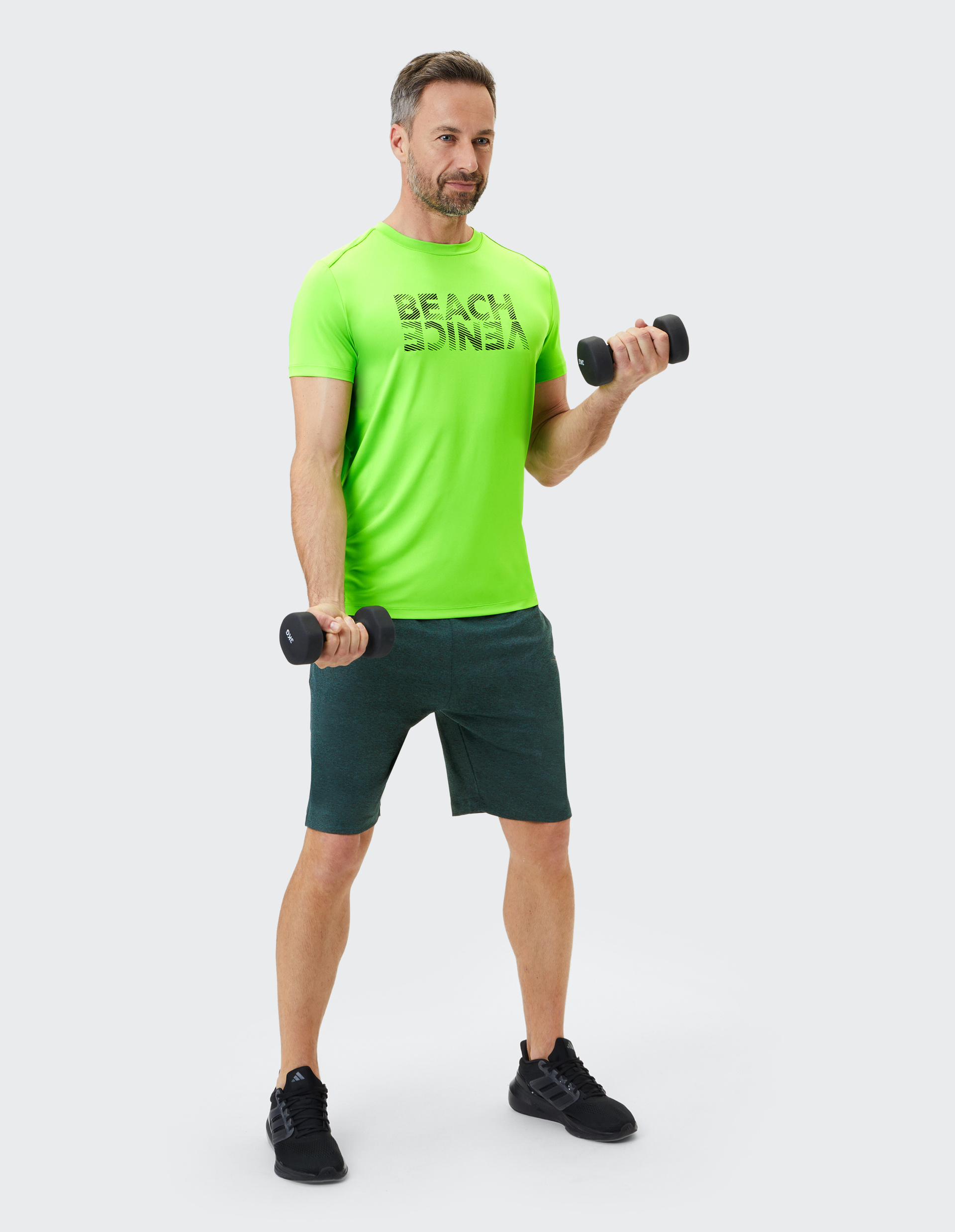Ein Mann in leuchtend grünem Shirt und dunklen Shorts hebt Hanteln, lächelnd, vor hellem Hintergrund, in sportlicher Pose.