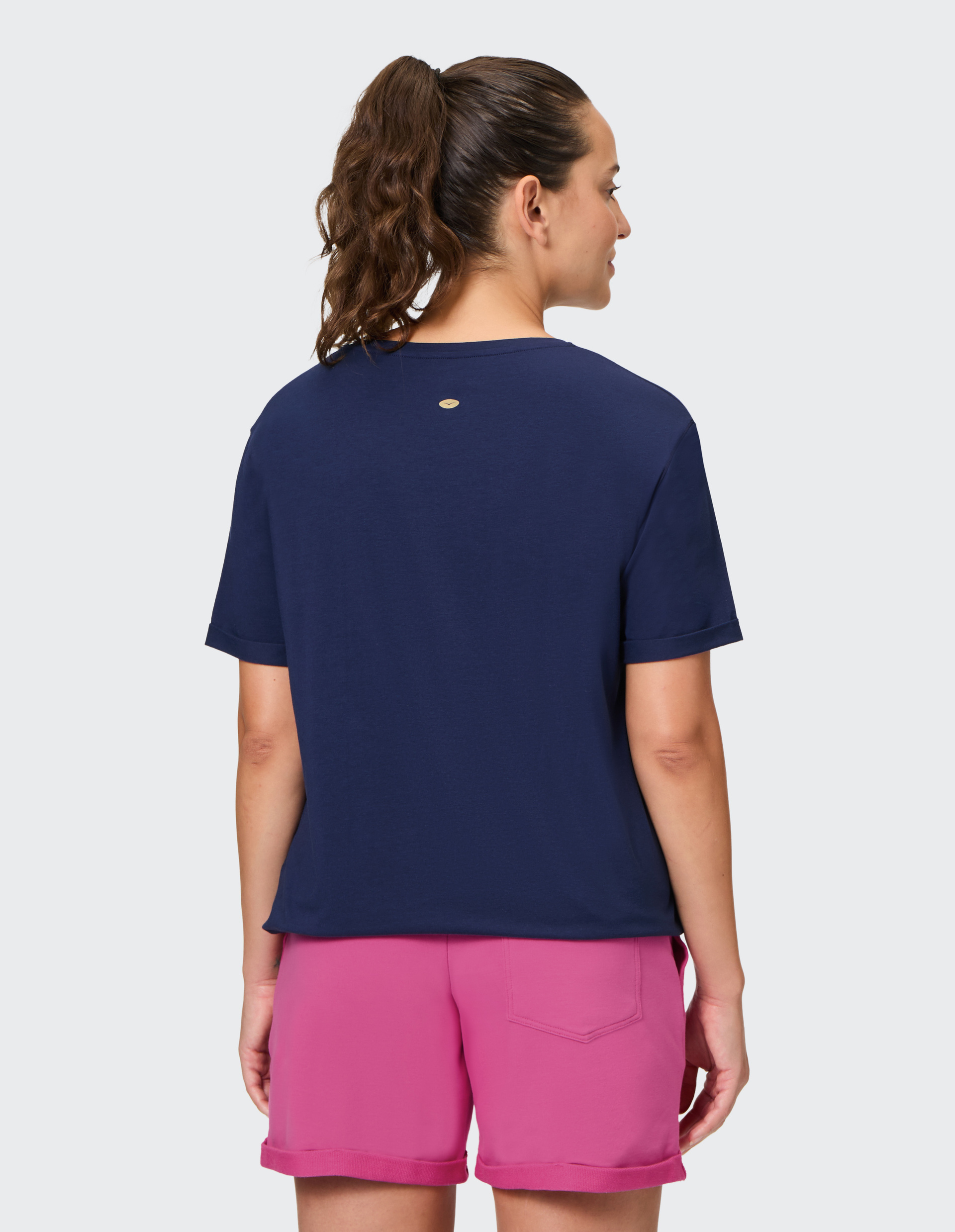 Frau mit lockigem Pferdeschwanz, trägt ein dunkelblaues T-Shirt und pinke Shorts, steht mit dem Rücken zur Kamera vor grauem Hintergrund.