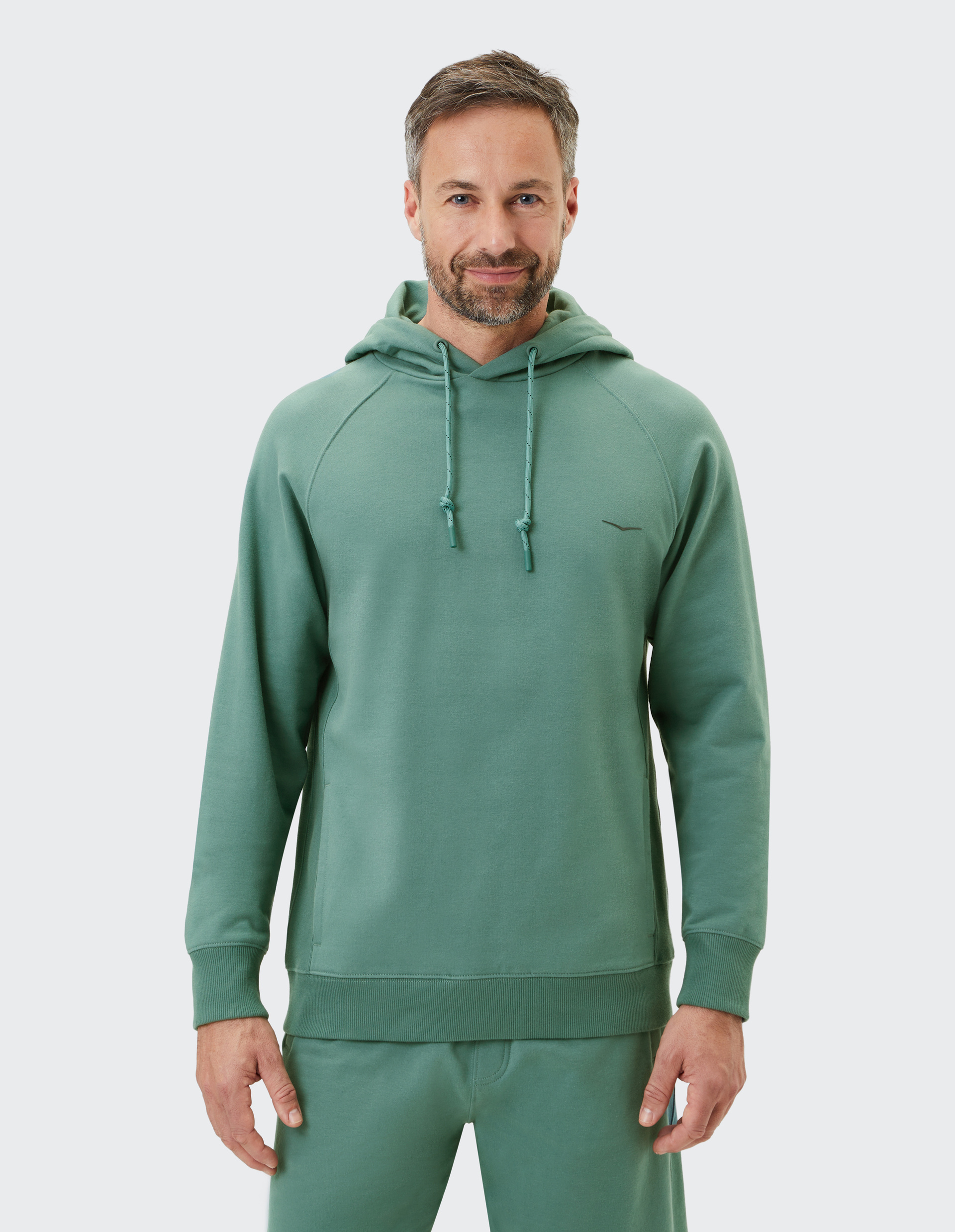 Ein lächelnder Mann trägt einen grünen Hoodie mit Kapuze und passenden Jogginghosen vor neutralem Hintergrund.