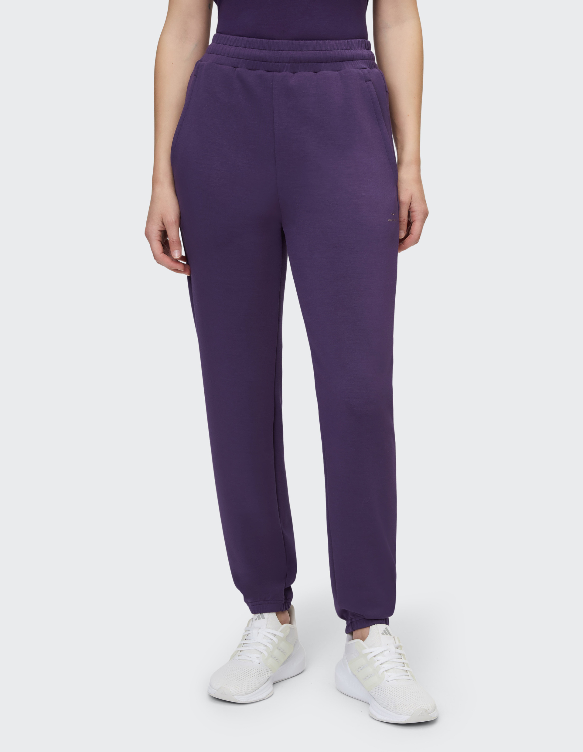 Person trägt lila Jogginghose mit elastischem Bund und weißen Sneakers, zeigt nur Unterkörper vor hellem Hintergrund.