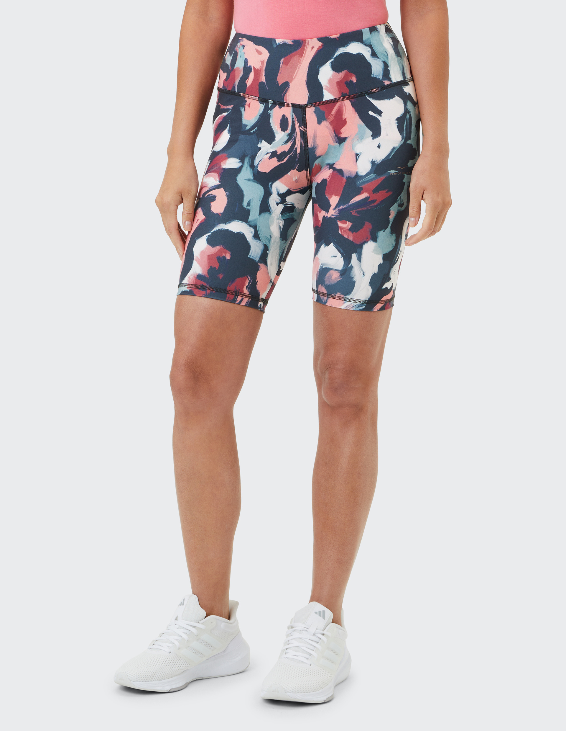 Frau trägt gemusterte, kniehohe Sportshorts mit rosa, marineblauen und weißen Akzenten, kombiniert mit weißen Turnschuhen.