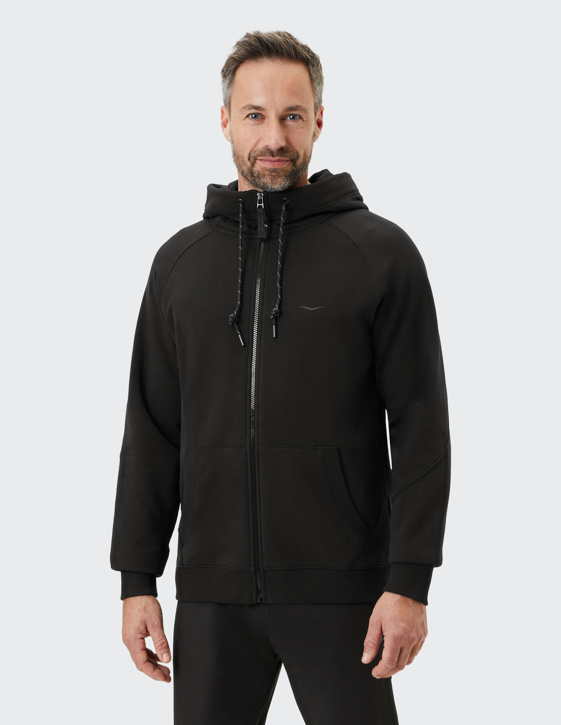 Ein Mann trägt eine schwarze Kapuzenjacke mit Reißverschluss und steht vor neutralem Hintergrund.