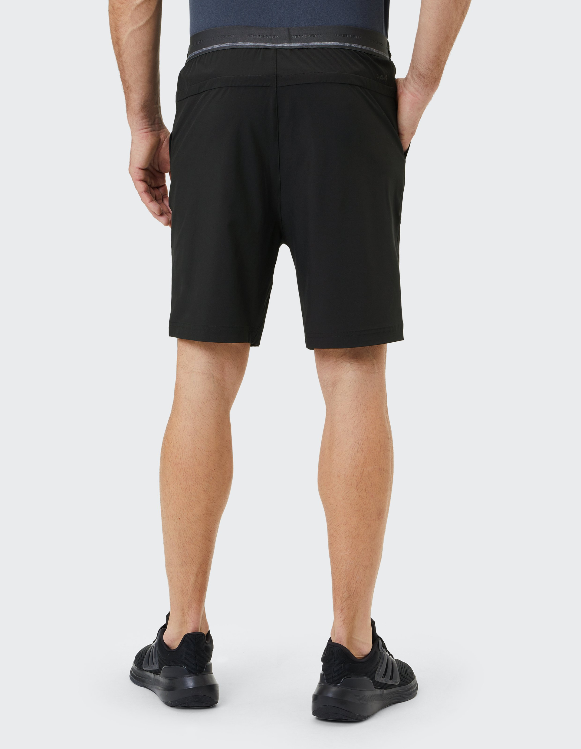 Rückansicht eines Menschen in schwarzer Sporthose und Sneakers, Hände in den Taschen, auf hellem Hintergrund stehend.