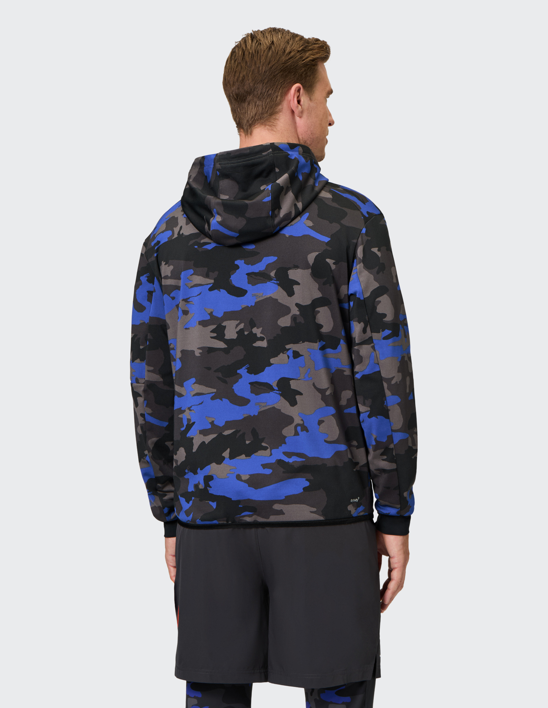 Mann trägt eine Kapuzenjacke mit blau-schwarzem Camouflage-Muster und dunkler Hose, Rückenansicht vor hellem Hintergrund.