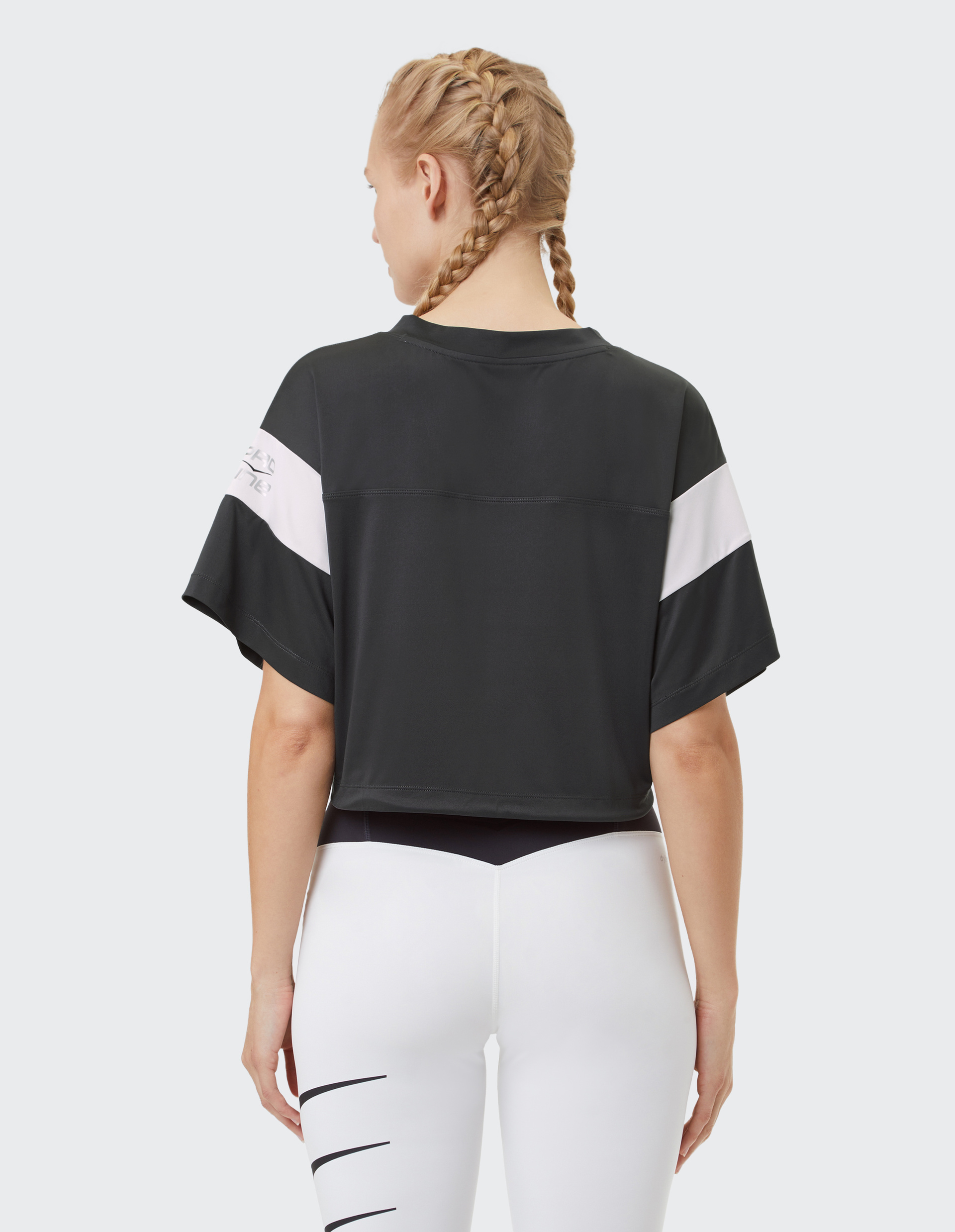 Frau mit zwei geflochtenen Zöpfen trägt ein schwarzes, kurzärmeliges Shirt und weiße Leggings mit schwarzen Streifen, von hinten gesehen.