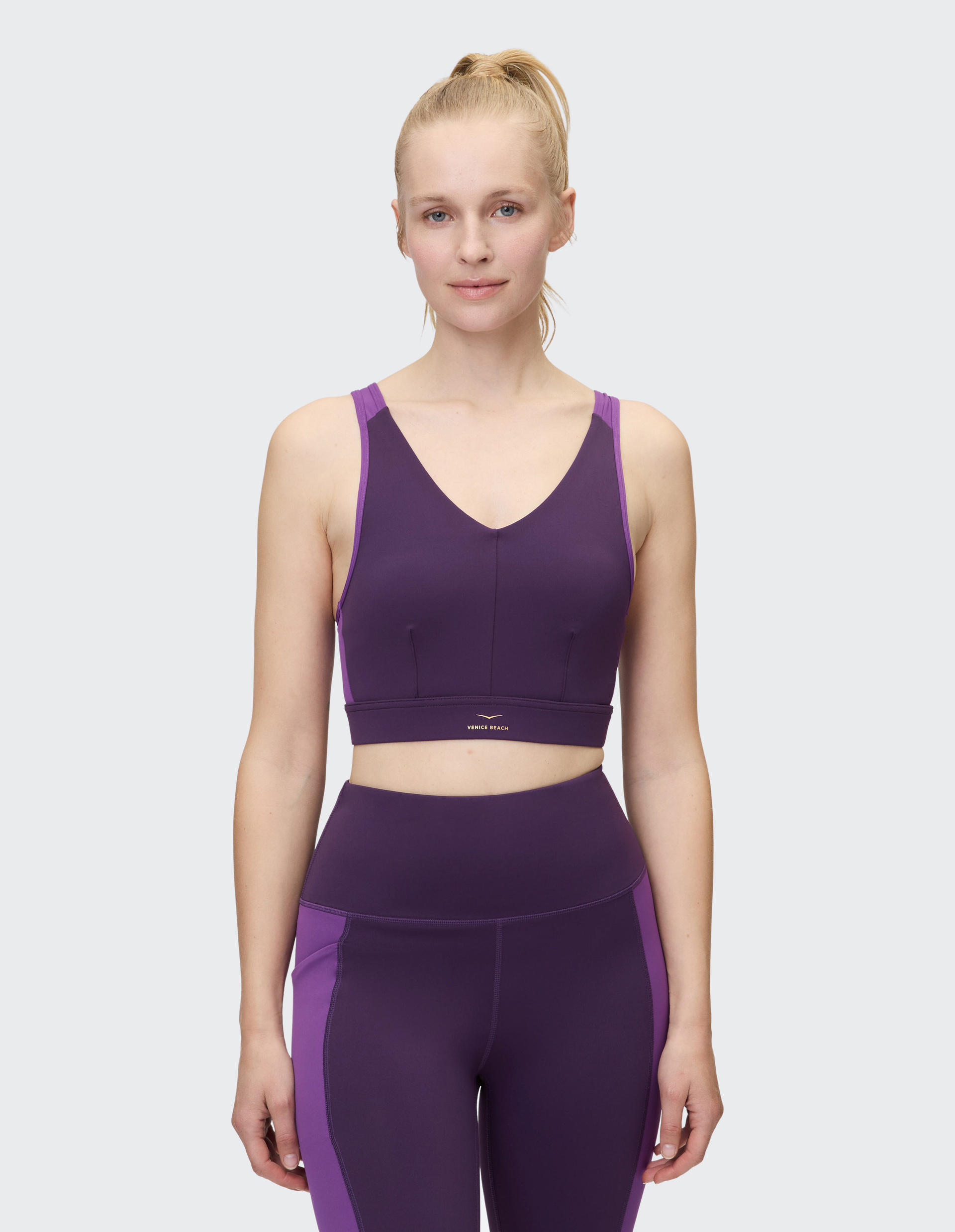 Frau mit blonden Haaren im Pferdeschwanz trägt lilafarbenes Sportoutfit mit V-Ausschnitt und lächelt dezent vor grauem Hintergrund.