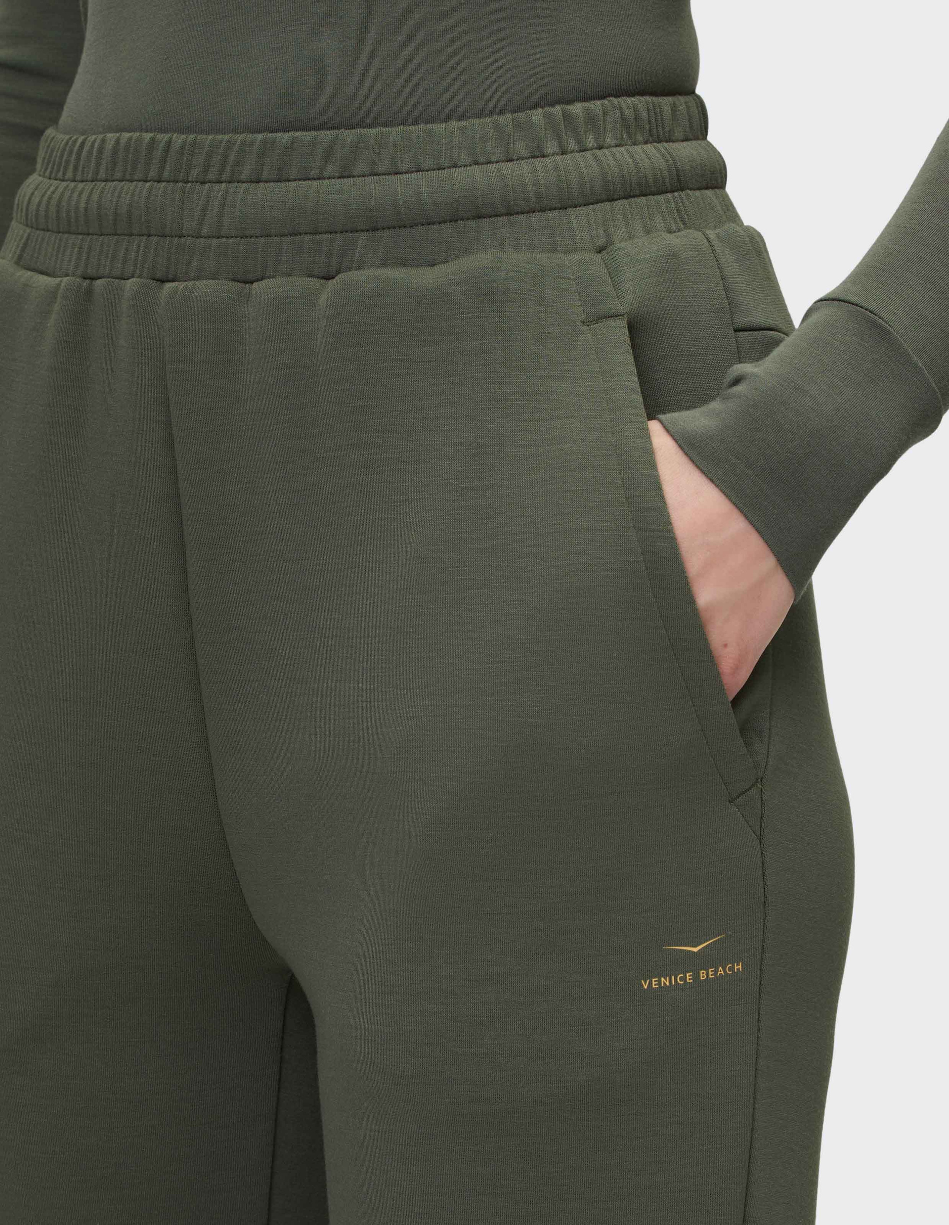 Dunkelgrüne Jogginghose mit elastischem Bund und Hand in der rechten Tasche, begleitet von einem kleinen Venice Beach Logo.