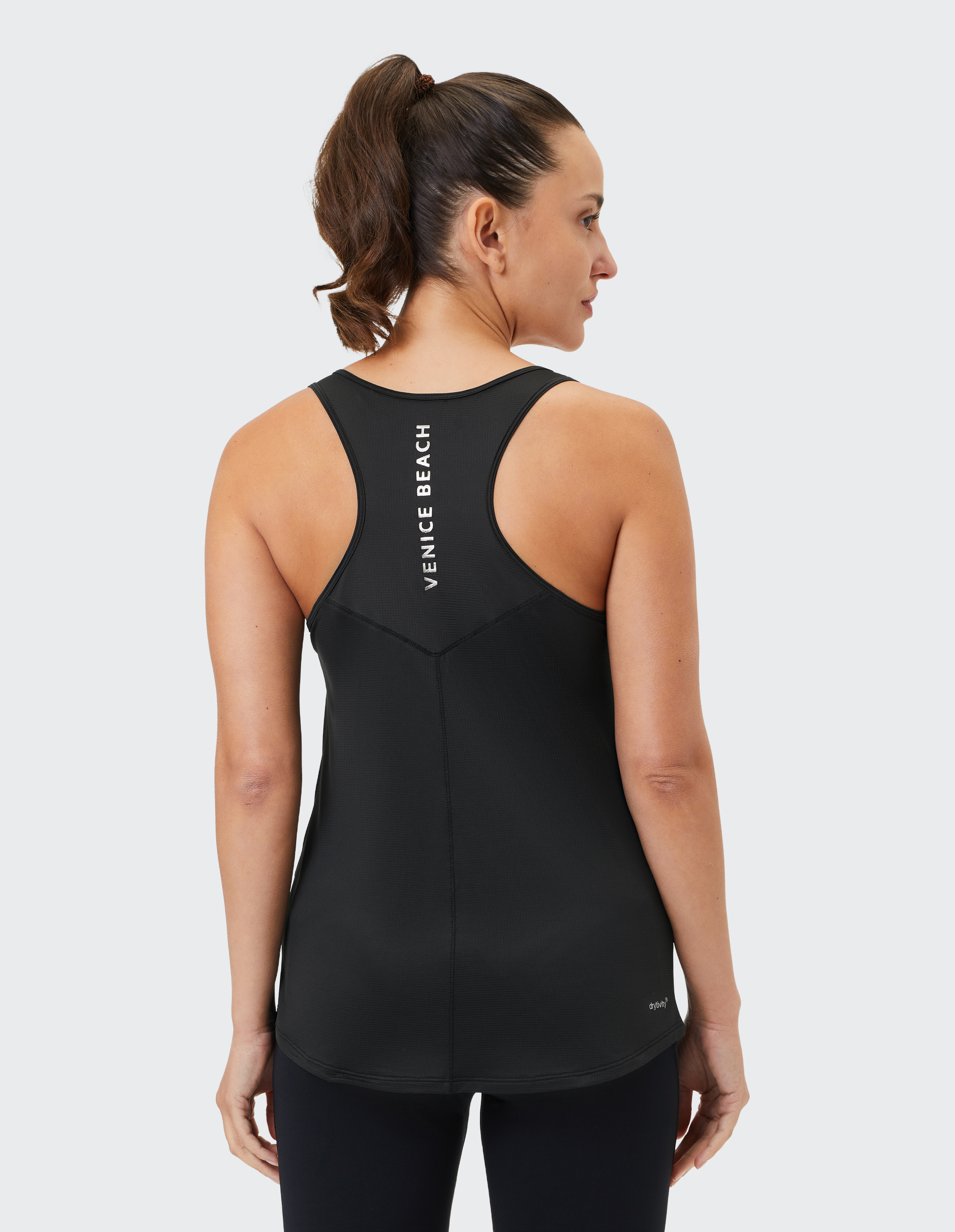 Frau mit Pferdeschwanz trägt schwarzes Tanktop mit "Venice Beach"-Schriftzug auf dem Rücken, steht in entspannter Haltung.