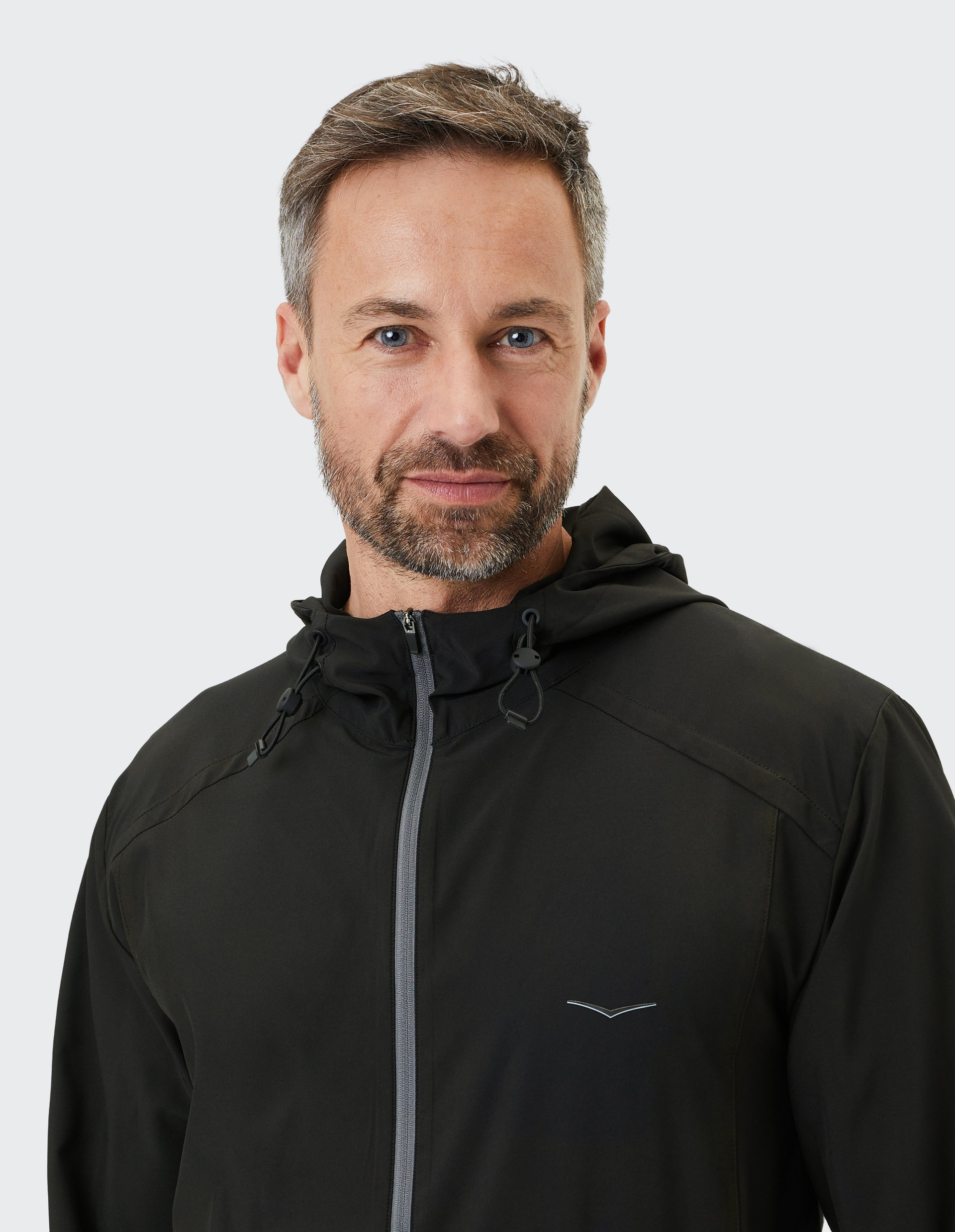 Ein Mann mit kurzem Haar und grauem Bart trägt eine schwarze Jacke mit Kapuze und Reißverschluss vor schlichtem, hellem Hintergrund.