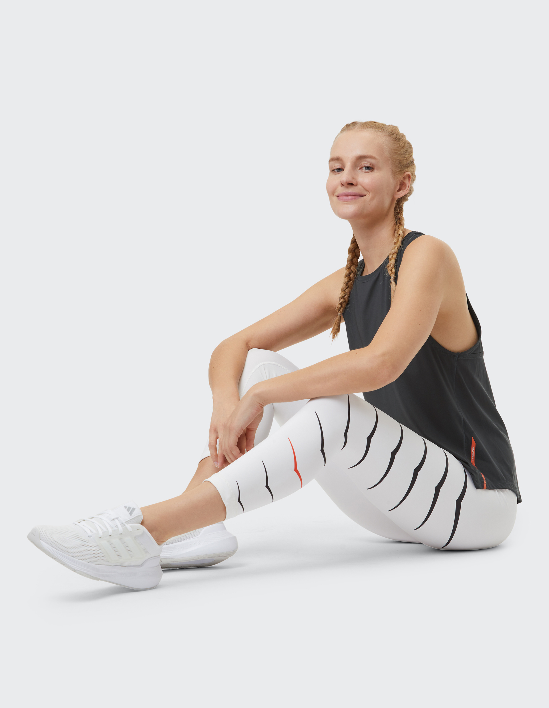 Frau mit Zöpfen sitzt lächelnd in Sportkleidung auf dem Boden, trägt weiße Leggings mit Muster und weiße Sneaker.