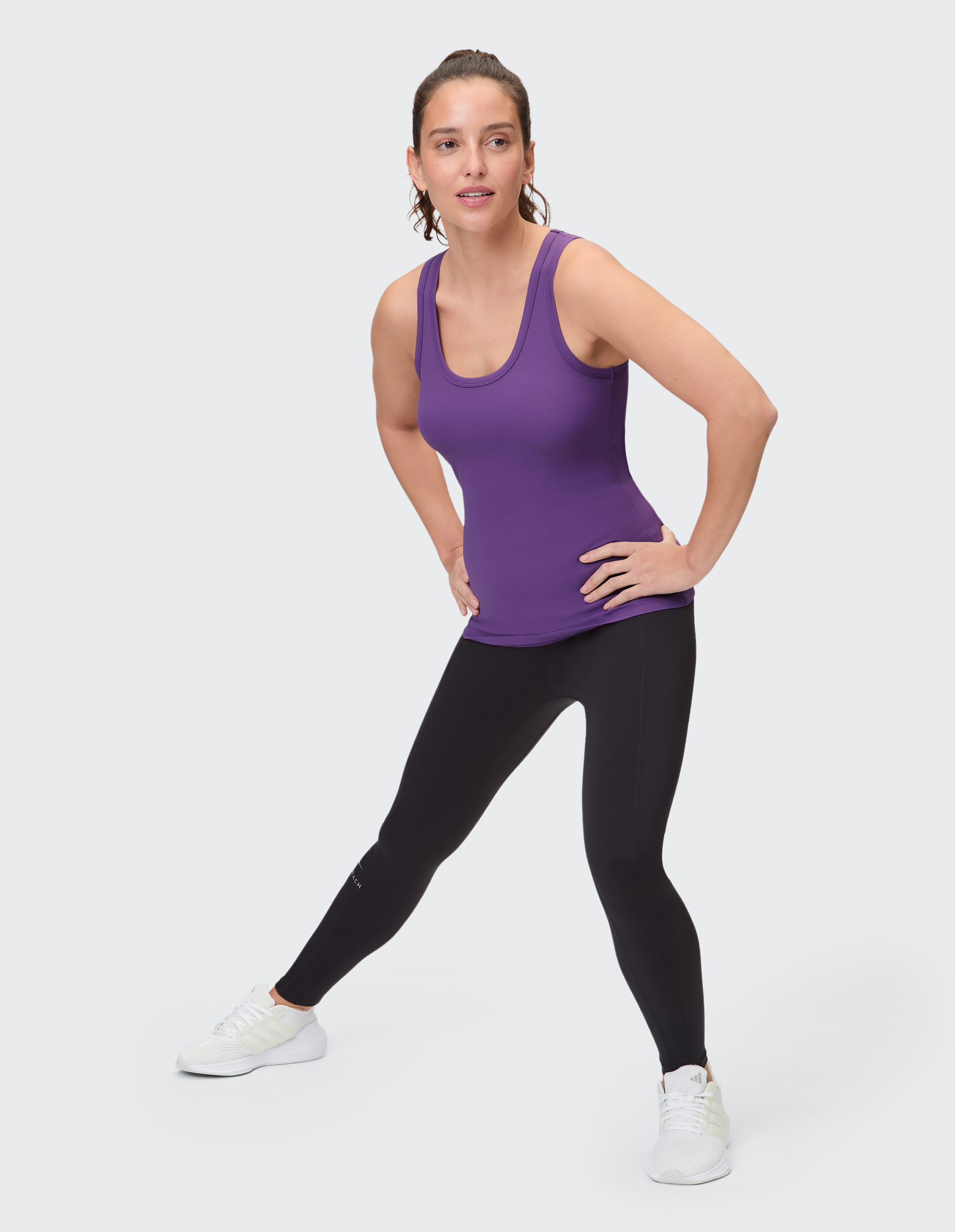 Frau in lila Tanktop und schwarzer Leggings steht aufrecht vor hellem Hintergrund, trägt weiße Turnschuhe und lächelt dezent.