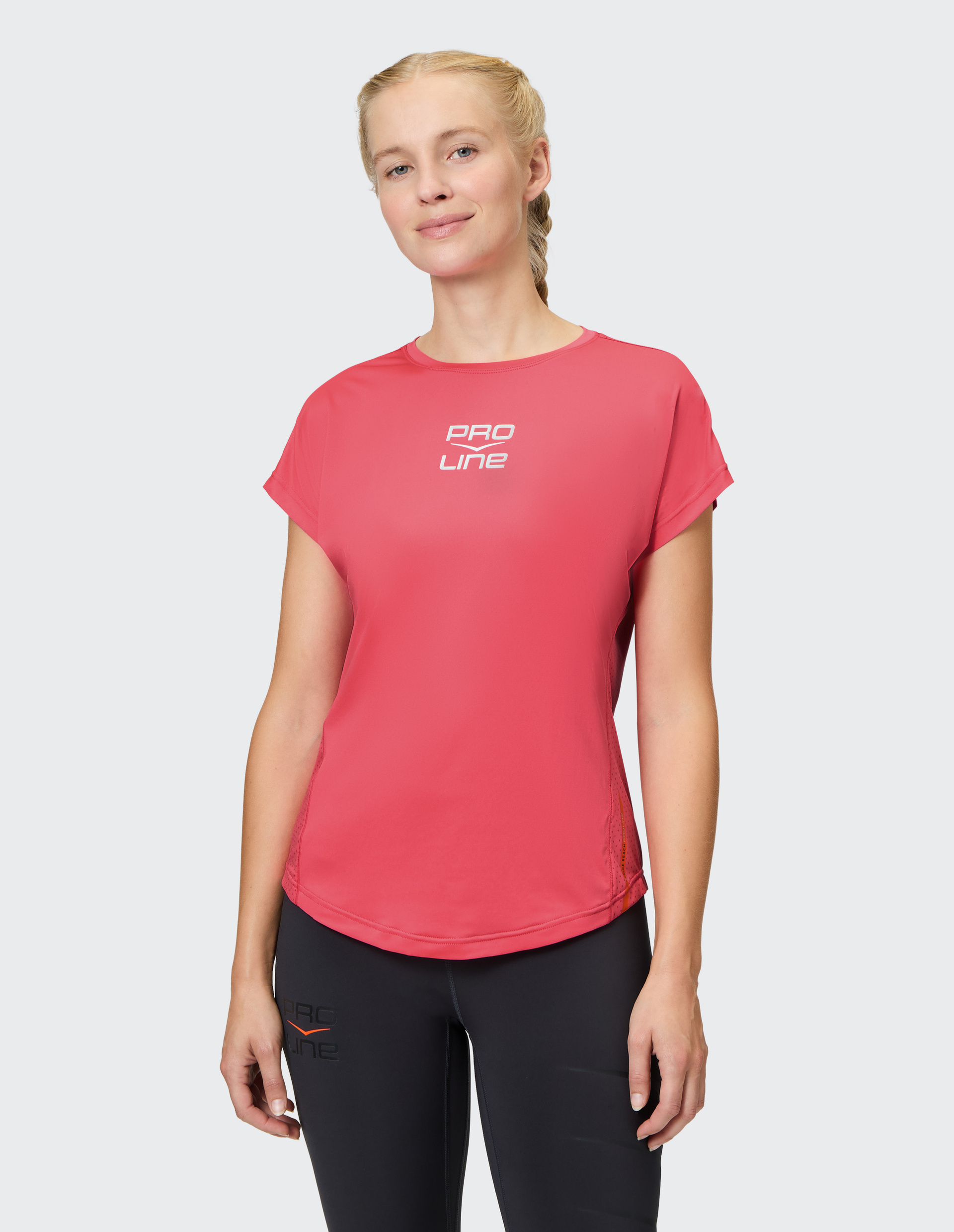 Person in rotem Pro Line T-Shirt und schwarzen Leggings mit seitlichem Blick, hell-blonde Haare geflochten, vor grauem Hintergrund.