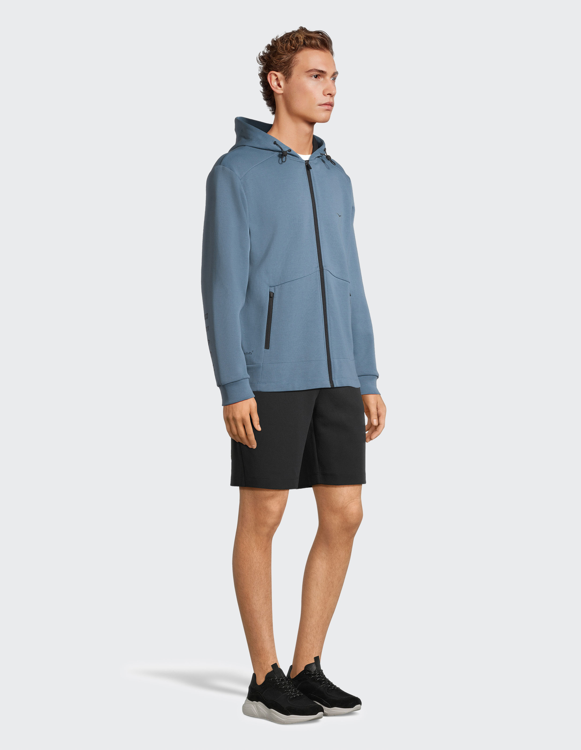 Ein Mann trägt eine blaue Kapuzenjacke, schwarze Shorts und Sneakers, steht seitlich vor einem weißen Hintergrund.