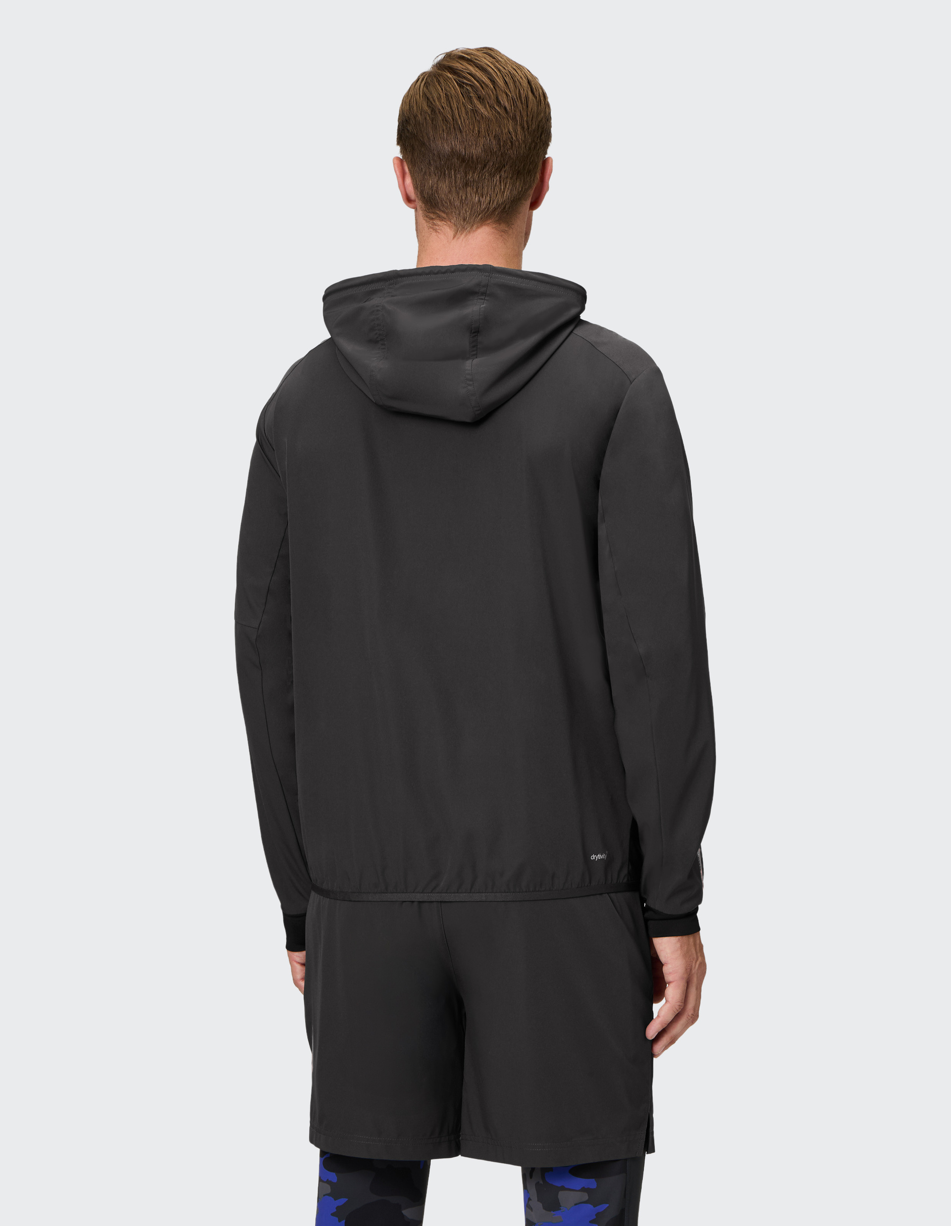 Ein Mann trägt eine schwarze Kapuzenjacke, schwarze Shorts und blaue Camouflage-Leggings, von hinten fotografiert.