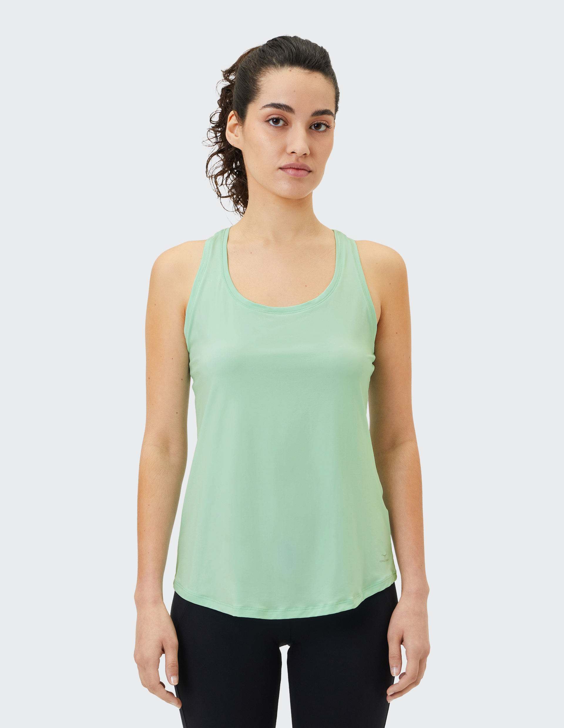 Frau mit lockigem Haar in Pferdeschwanz trägt ein mintgrünes ärmelloses Top und schwarze Leggings vor neutralem Hintergrund.